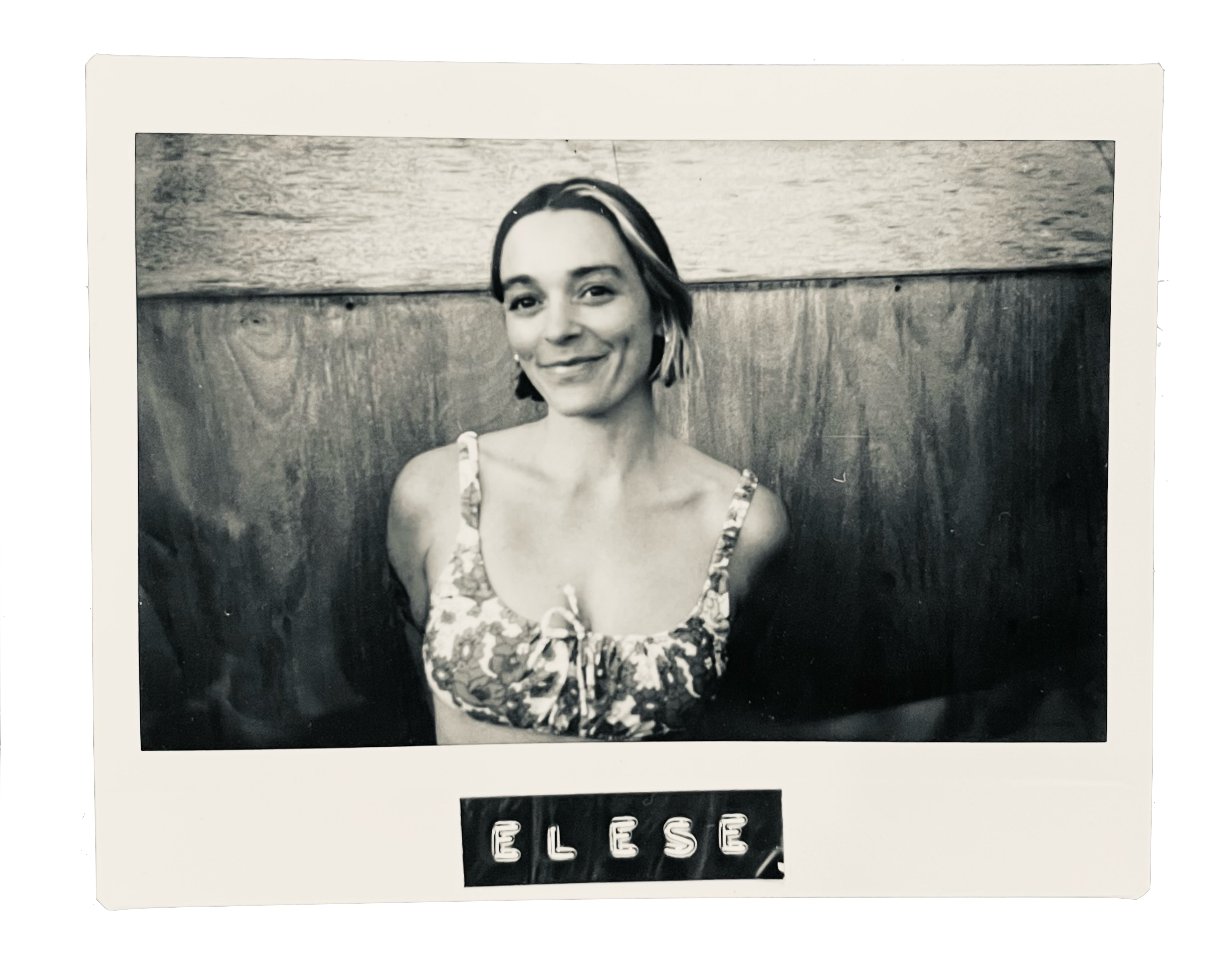 Black and white photo of a woman with short hair in a tank top, sitting in front of a wooden wall, smiling, with the name 'ELISE' displayed below.