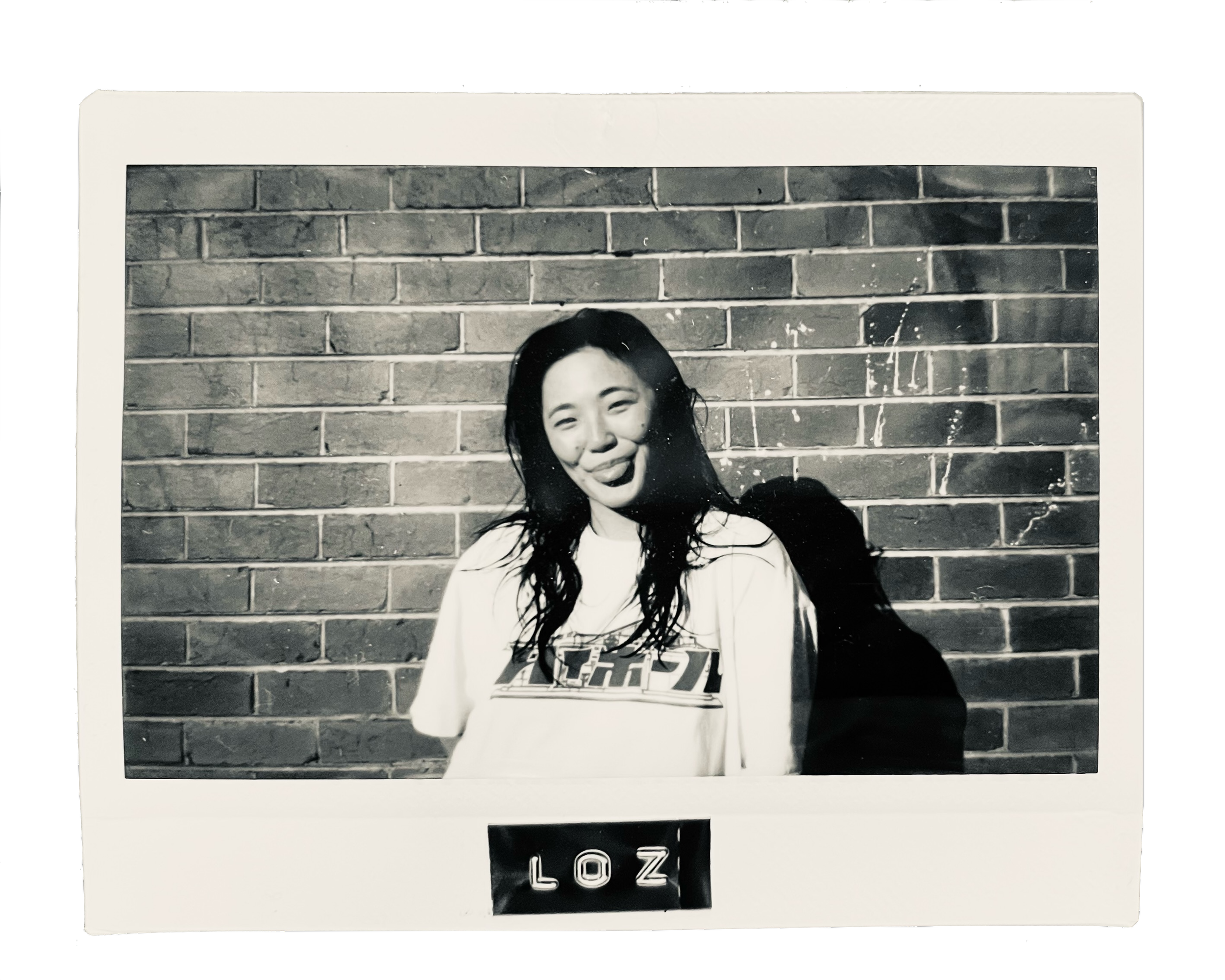 A black-and-white photo of a smiling woman with wet hair, standing in front of a brick wall. The photo appears to be a Polaroid with the word "LOZ" at the bottom.