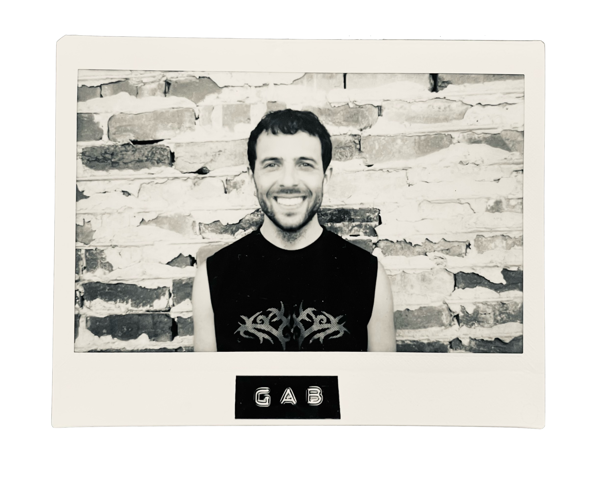 Black-and-white photo of a smiling man with a beard and short dark hair, wearing a sleeveless shirt with a tribal design, standing in front of a brick wall.