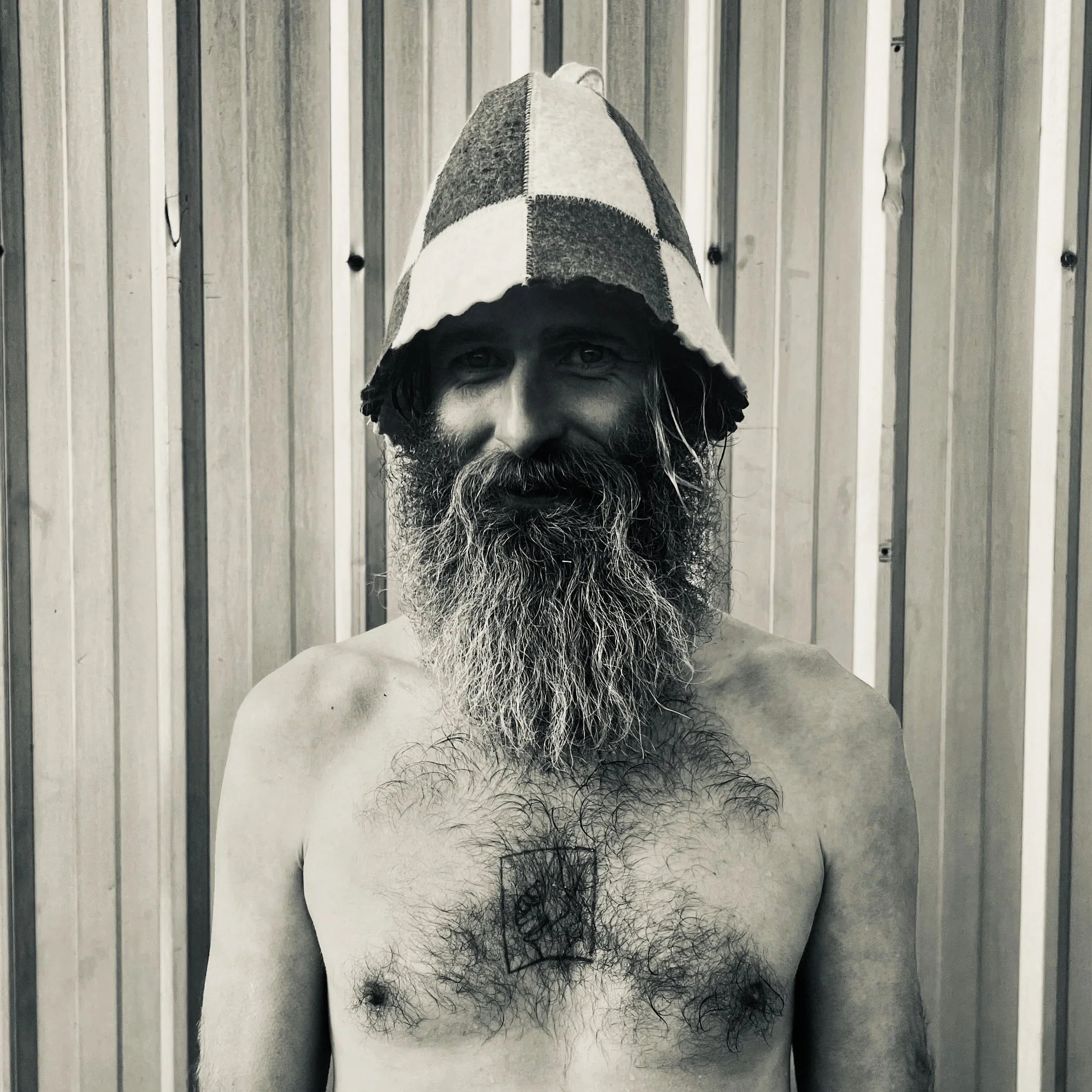 Black and white photo of a shirtless man with a long beard, wearing a hooded cap, standing in front of a wooden panel wall.