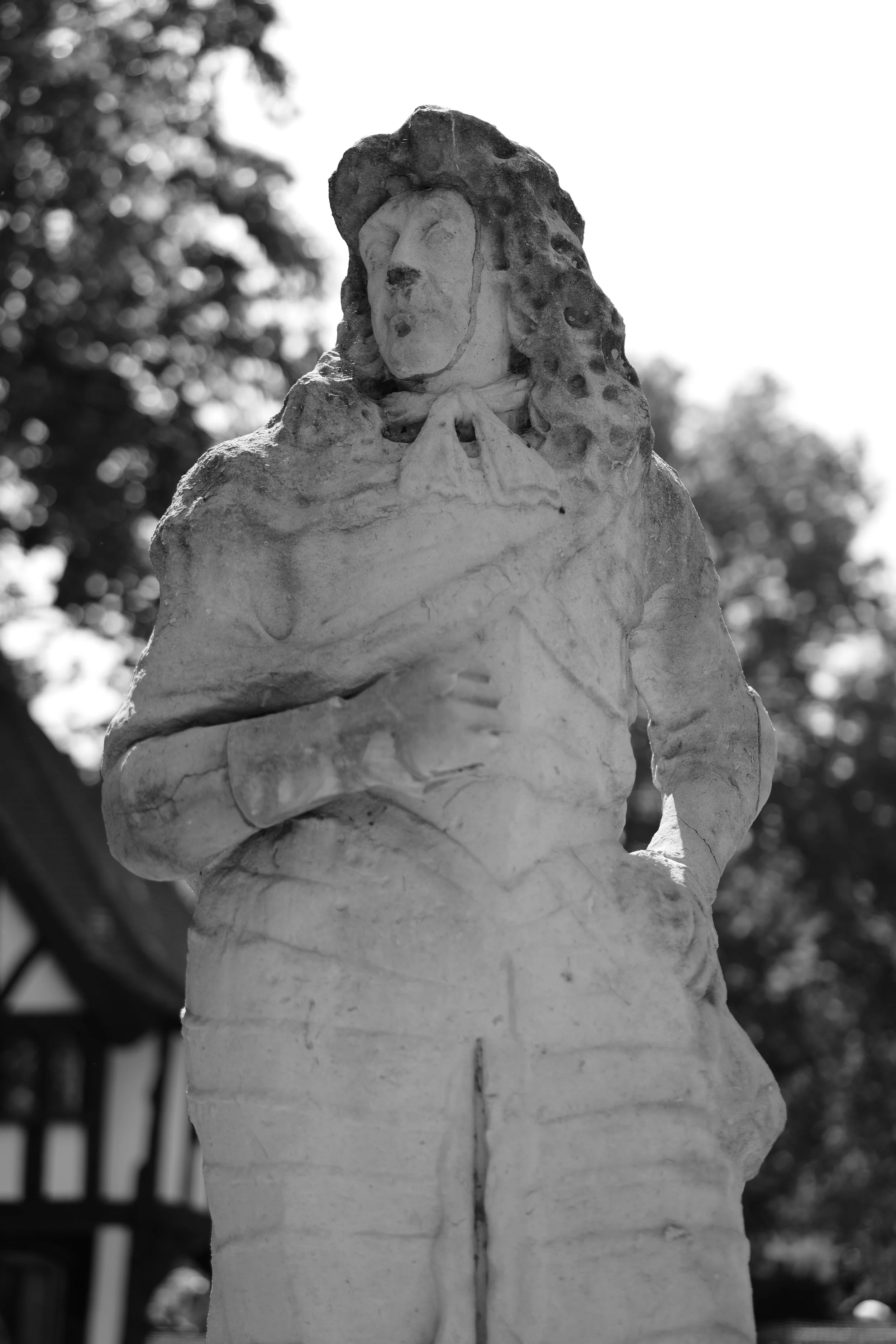 A black and white photograph of a statue of Jesus Christ wearing a crown of thorns, with a cloth draped over his shoulders and arms crossed in front of his chest, outdoors with blurred trees in the background.