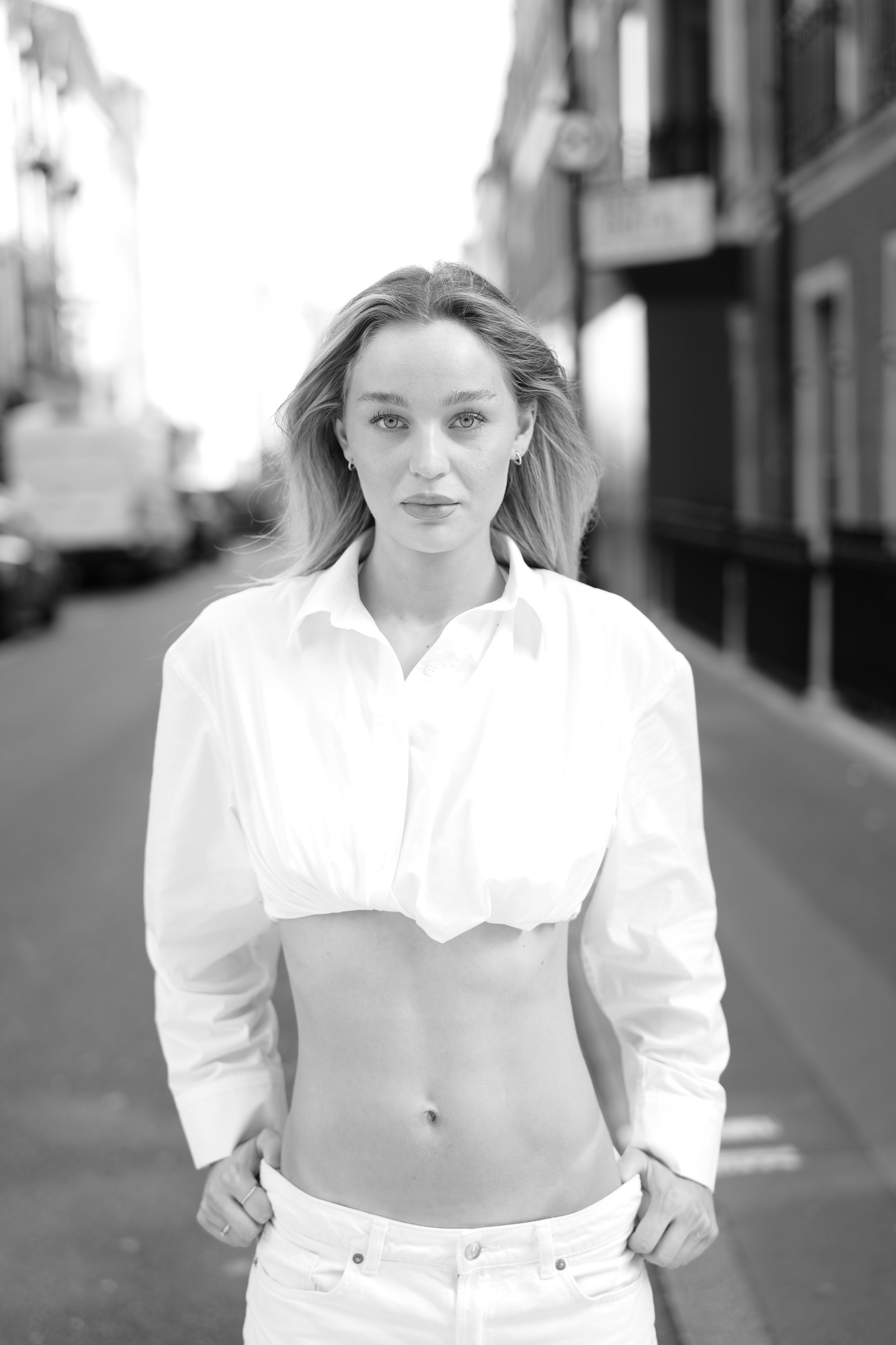 A black and white photo of a young woman with light eyes and long hair, standing outdoors in an urban setting. She wears a white shirt with the top buttons undone, exposing her midriff, and has her hands on her hips.