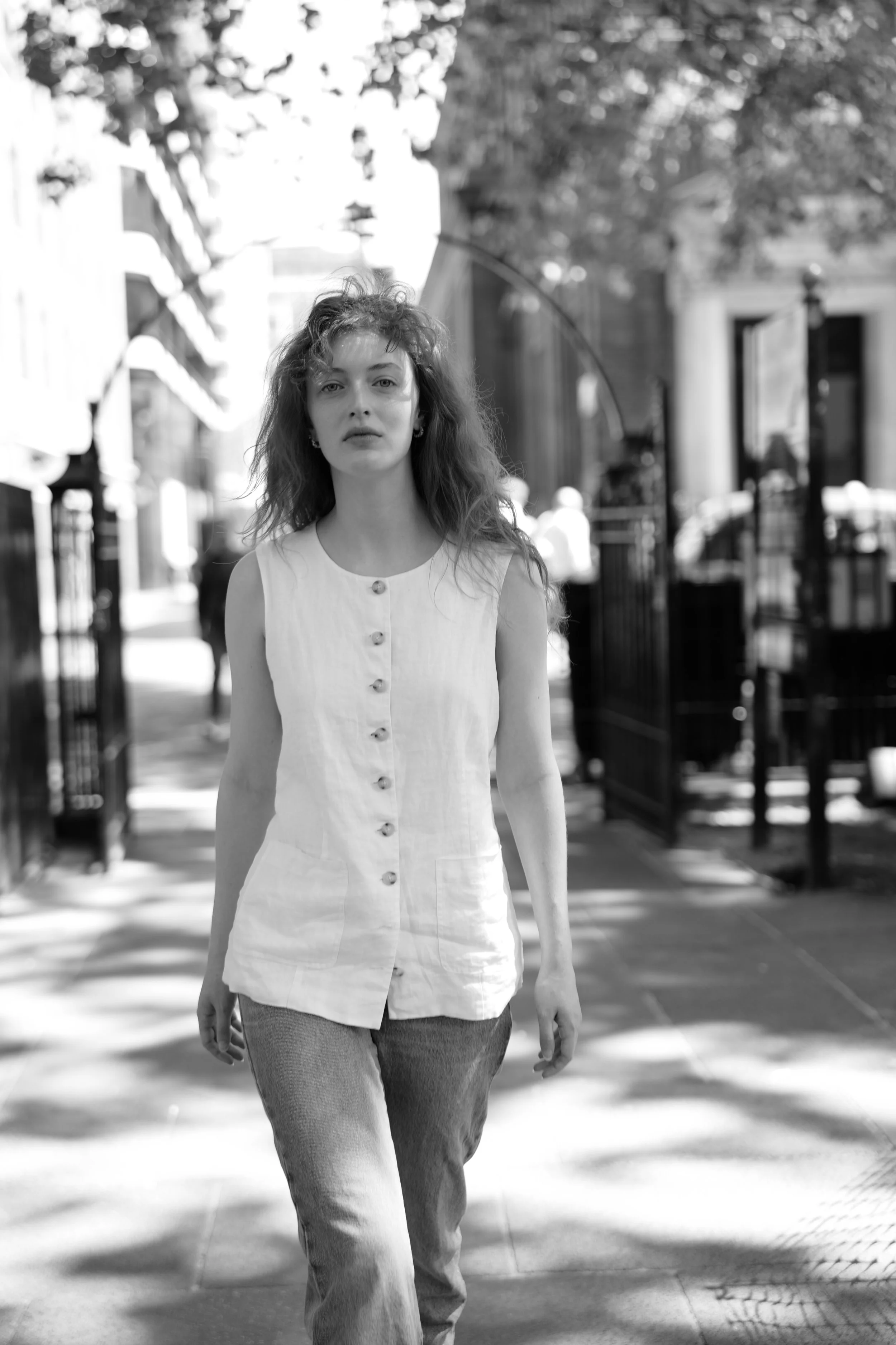 A woman walking outdoors in daytime, wearing a sleeveless buttoned top and jeans, with blurred trees and buildings in the background.