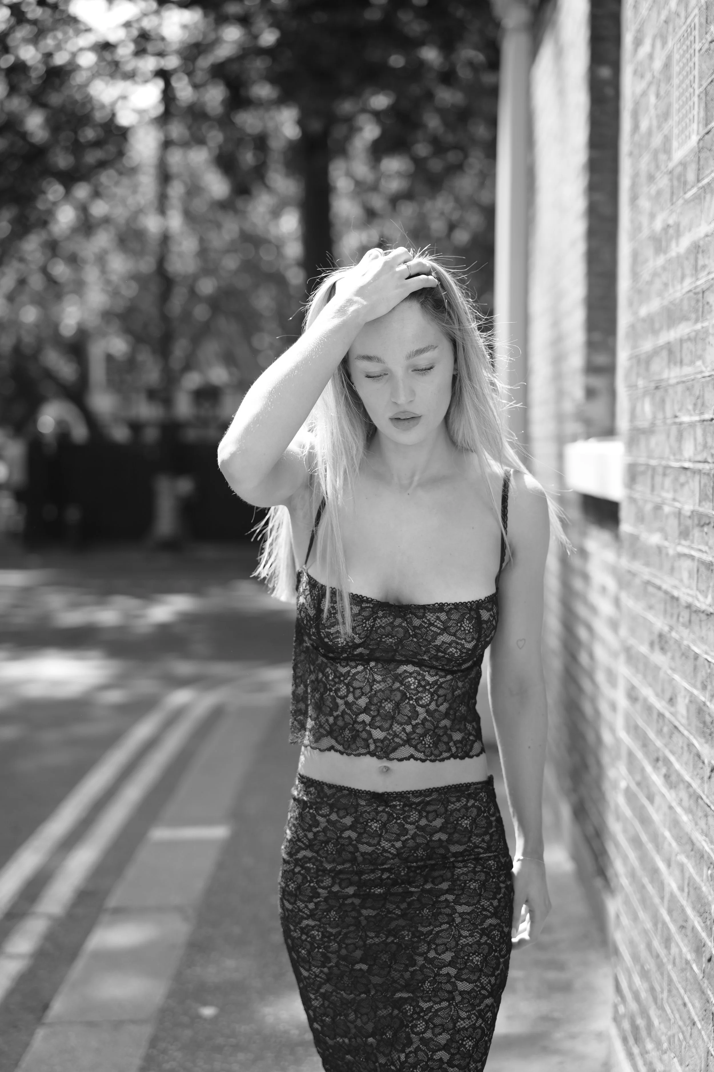 A young woman with long hair wearing a black lace dress stands outdoors near a brick wall, with her eyes closed and her hand resting on her head, sunlit trees in the background.