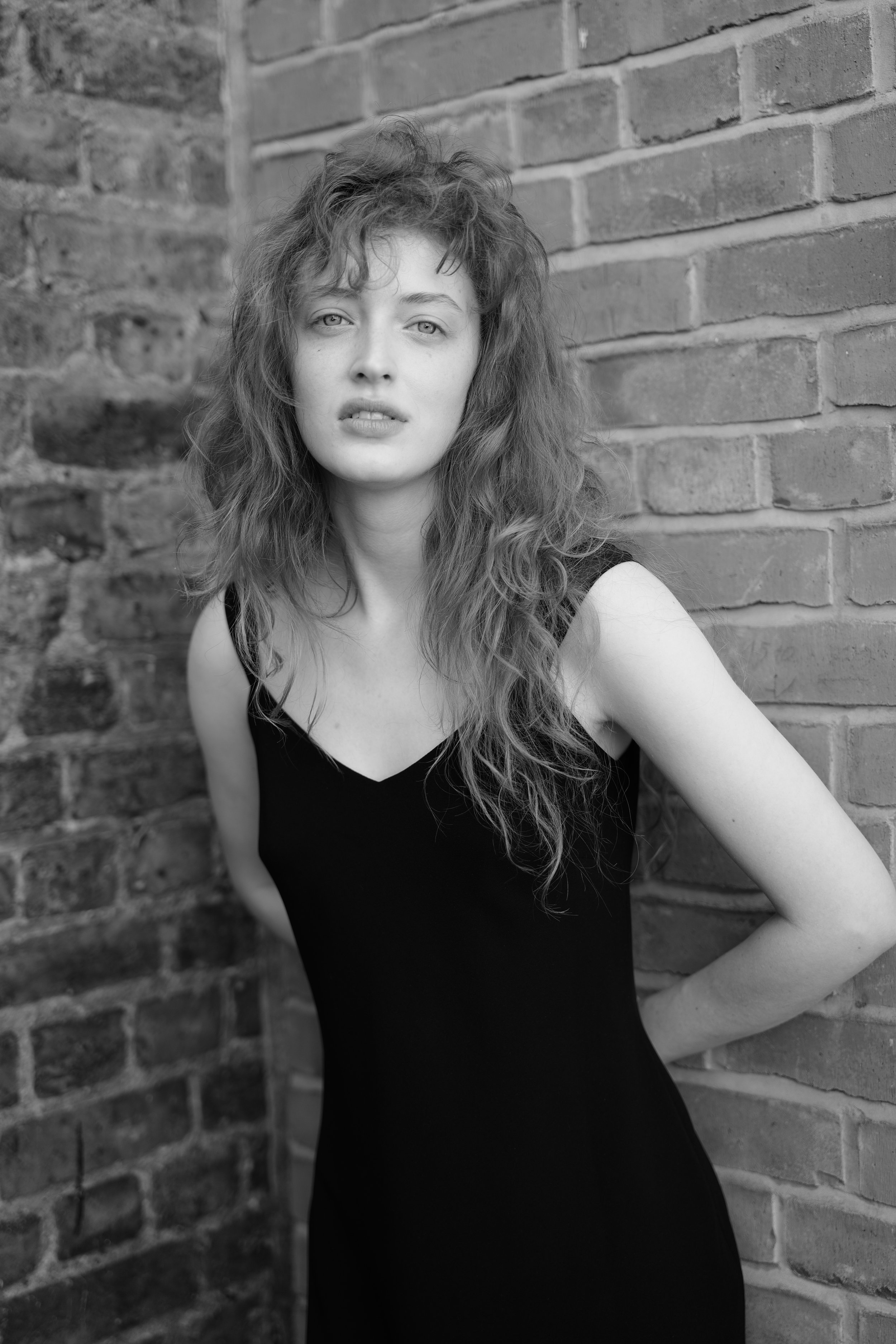 A black-and-white photo of a young woman with curly hair, wearing a sleeveless black top, leaning against a brick wall and looking at the camera.