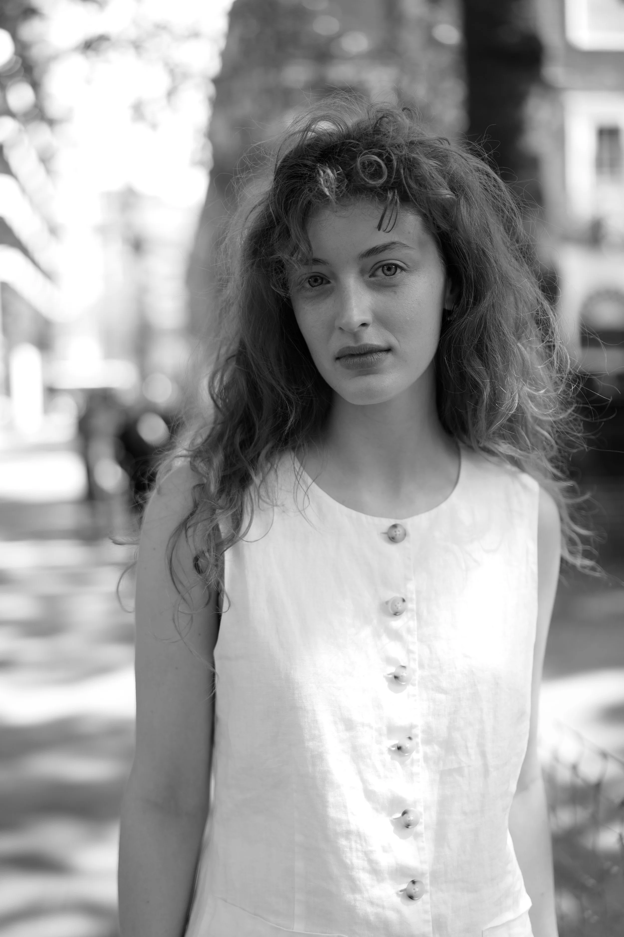 Black and white photo of a woman with curly hair wearing a sleeveless dress with buttons down the front, standing outdoors.