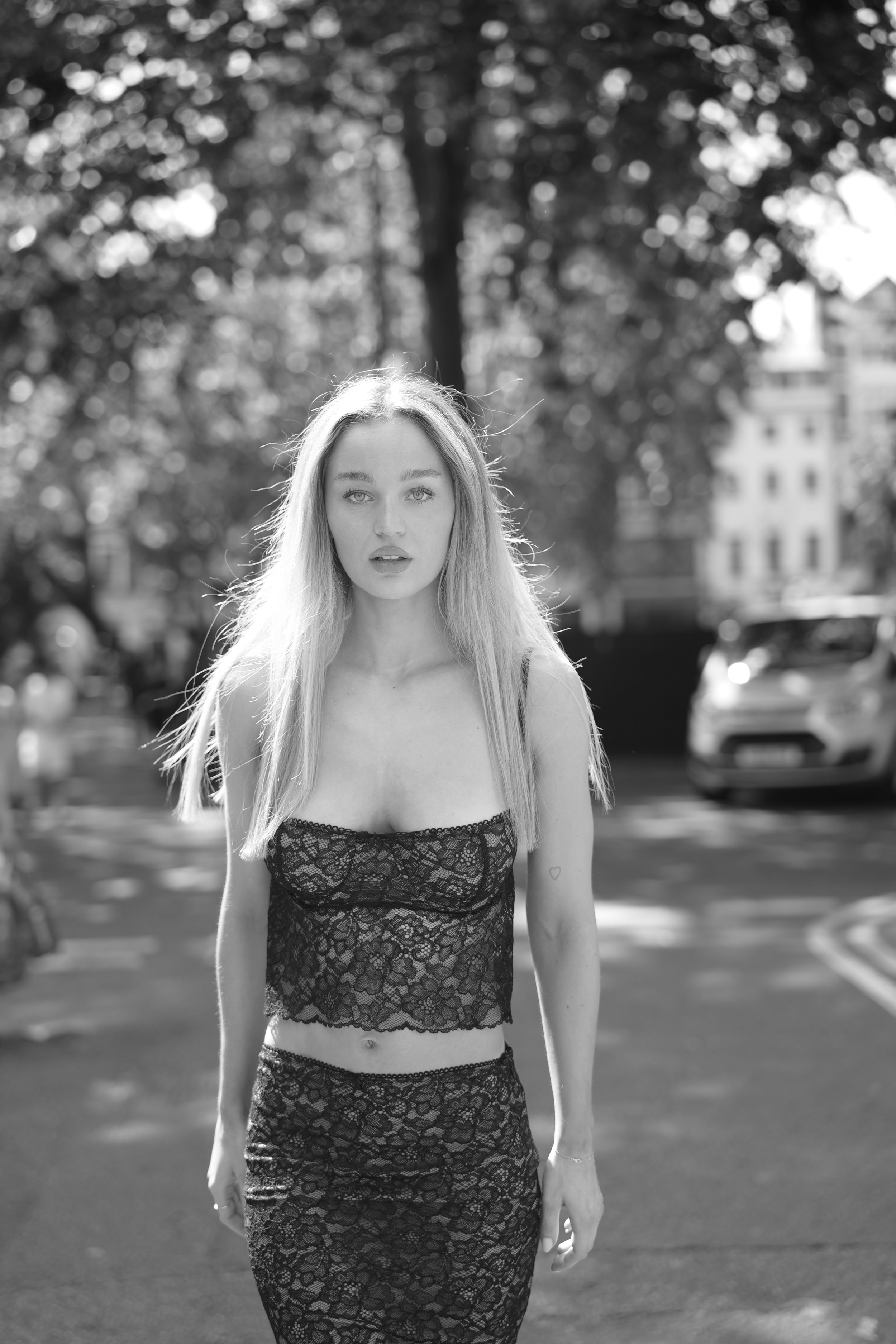 A black and white photograph of a young woman with long, light-colored hair standing on a street. She is wearing a lace crop top and matching skirt, and looking directly at the camera with a neutral expression. The background features trees and blurr