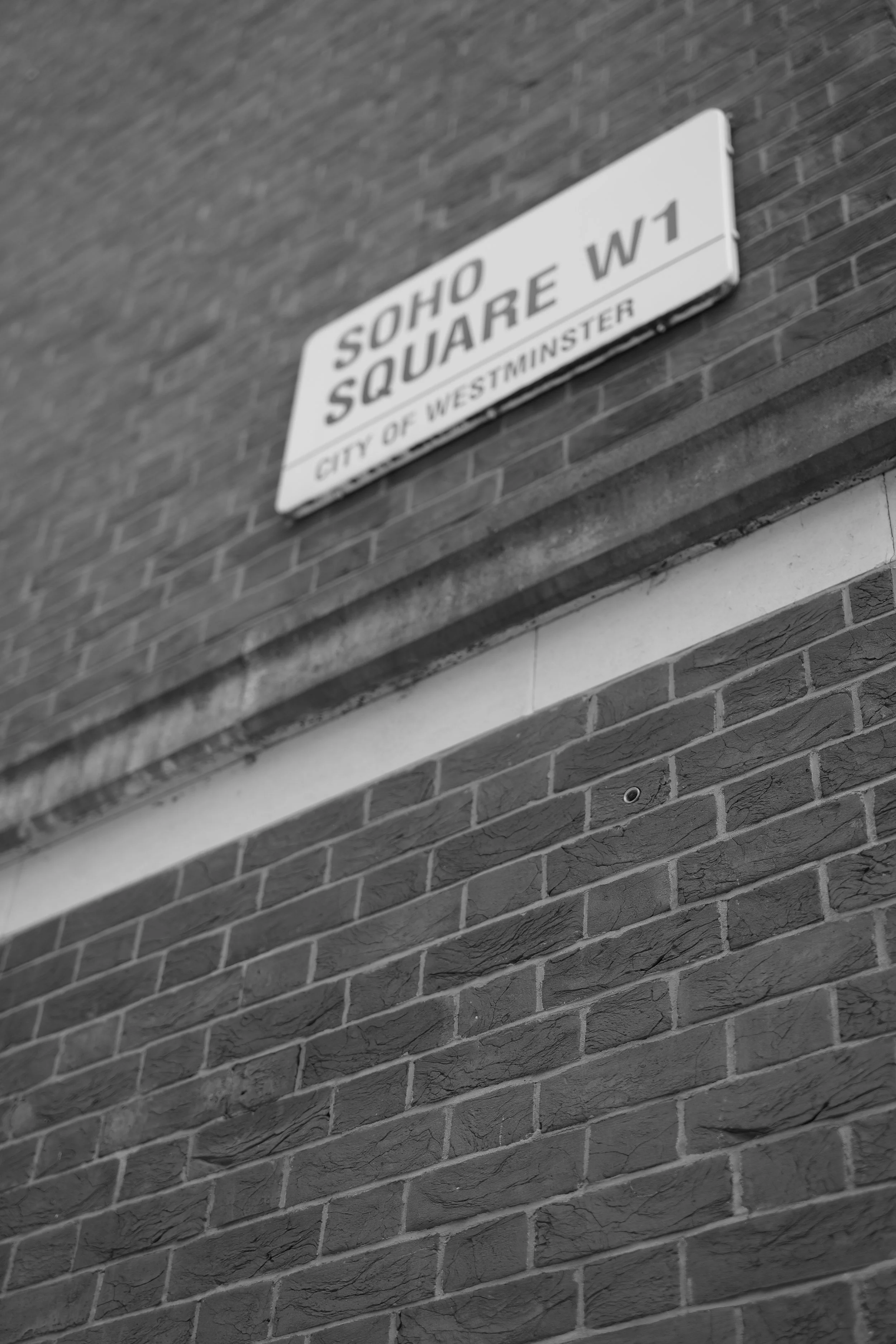 Street sign reading 'SOHO SQUARE' mounted on a brick wall, with smaller text 'CITY OF WESTMINSTER' below.