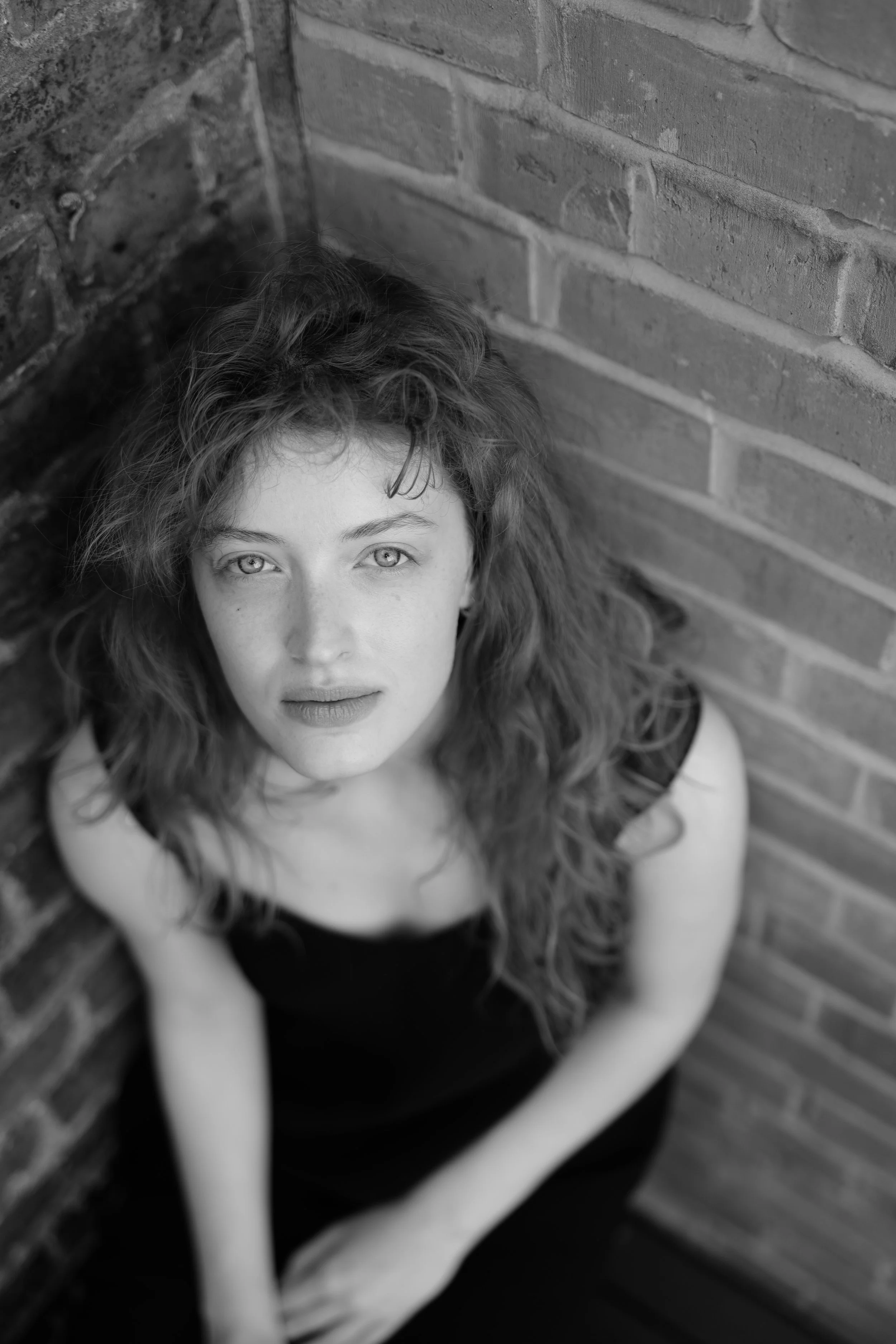 A woman with curly hair and light-colored eyes, sitting against a brick wall, looking directly at the camera with a neutral expression.