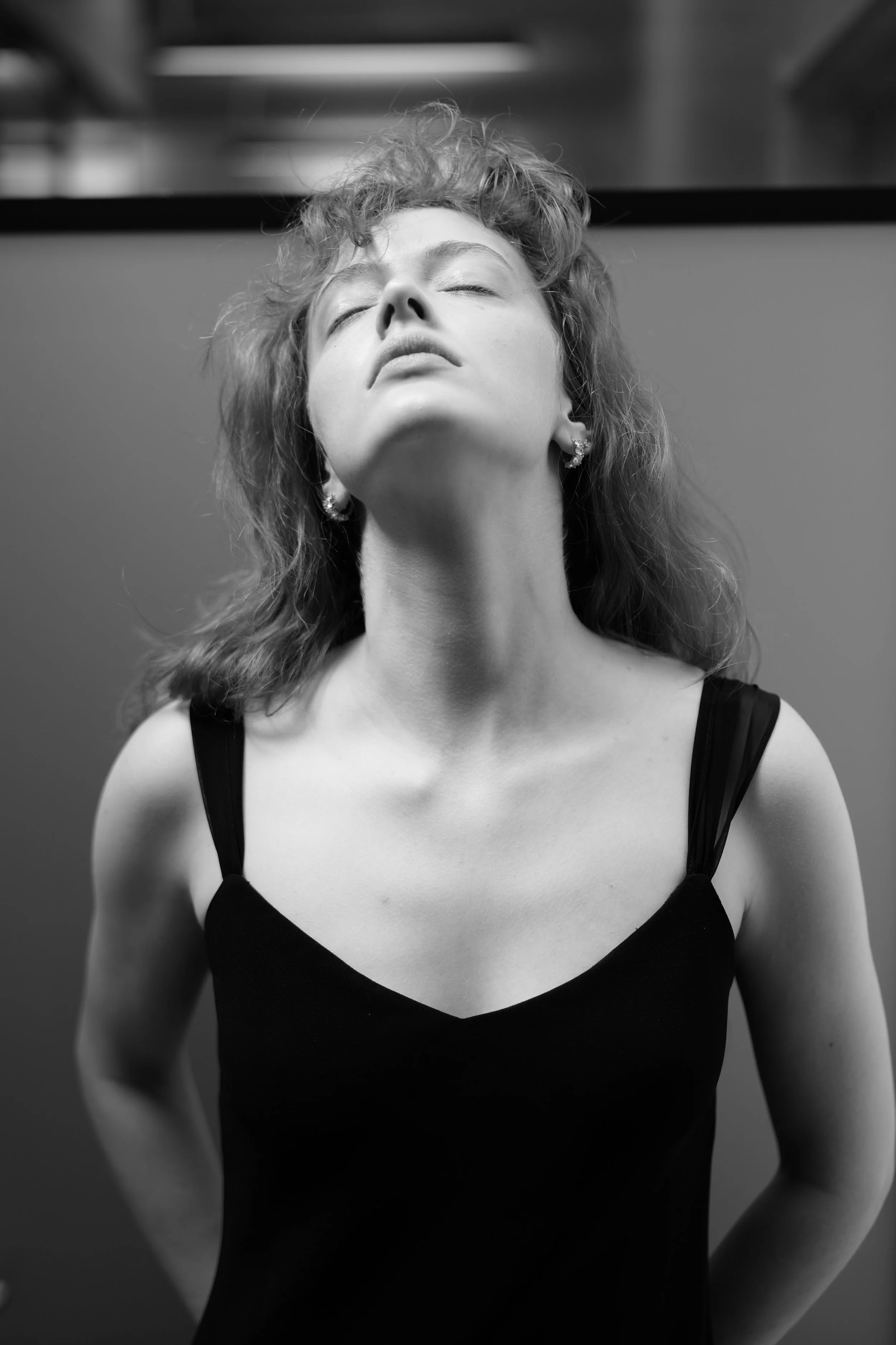 Black and white photograph of a woman with curly hair and closed eyes, wearing a black top with thin straps and earrings.