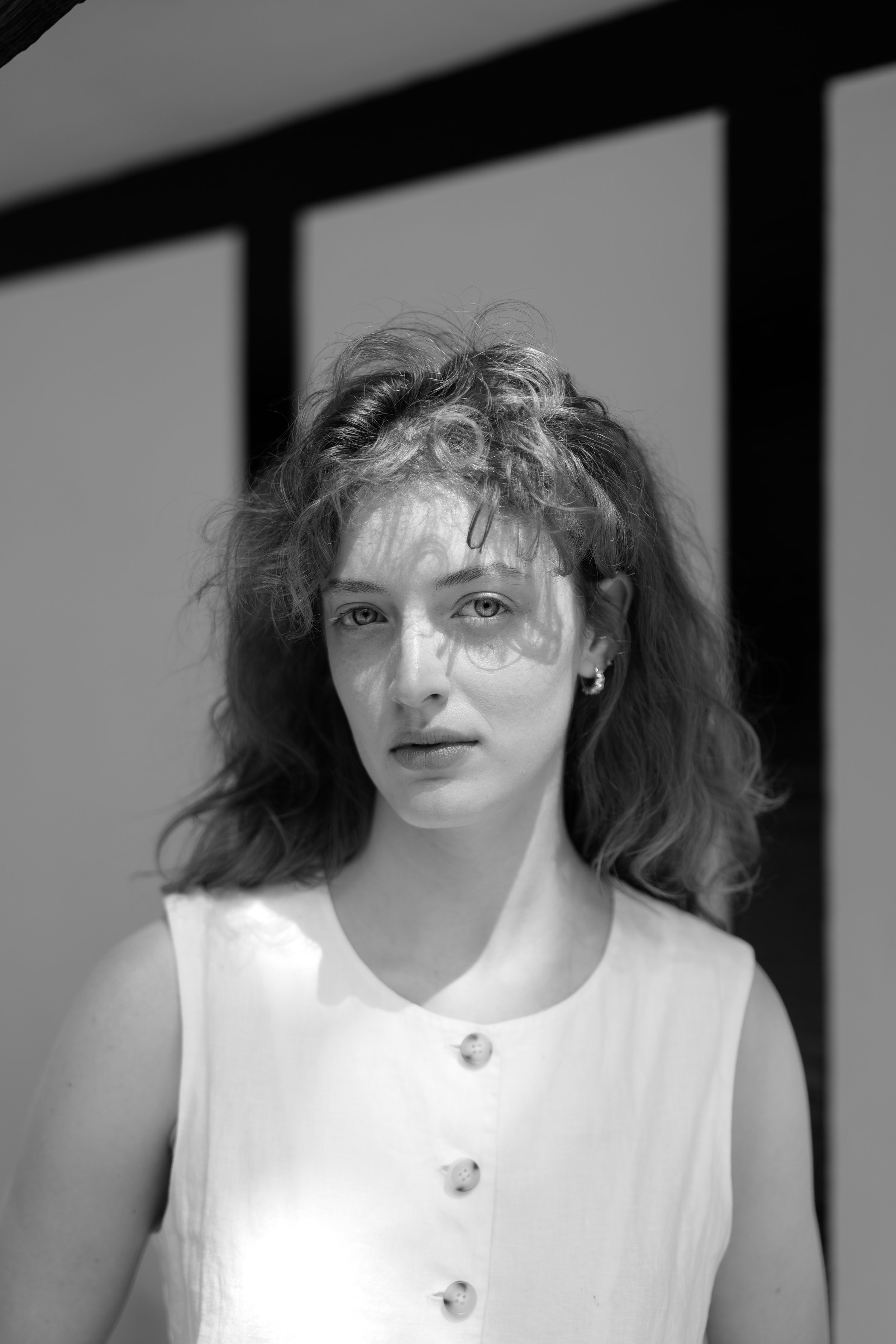 Black and white photo of a woman with curly hair, wearing a sleeveless top with buttons, looking directly at the camera, with a shadow of a grid pattern cast on her face and shoulders.