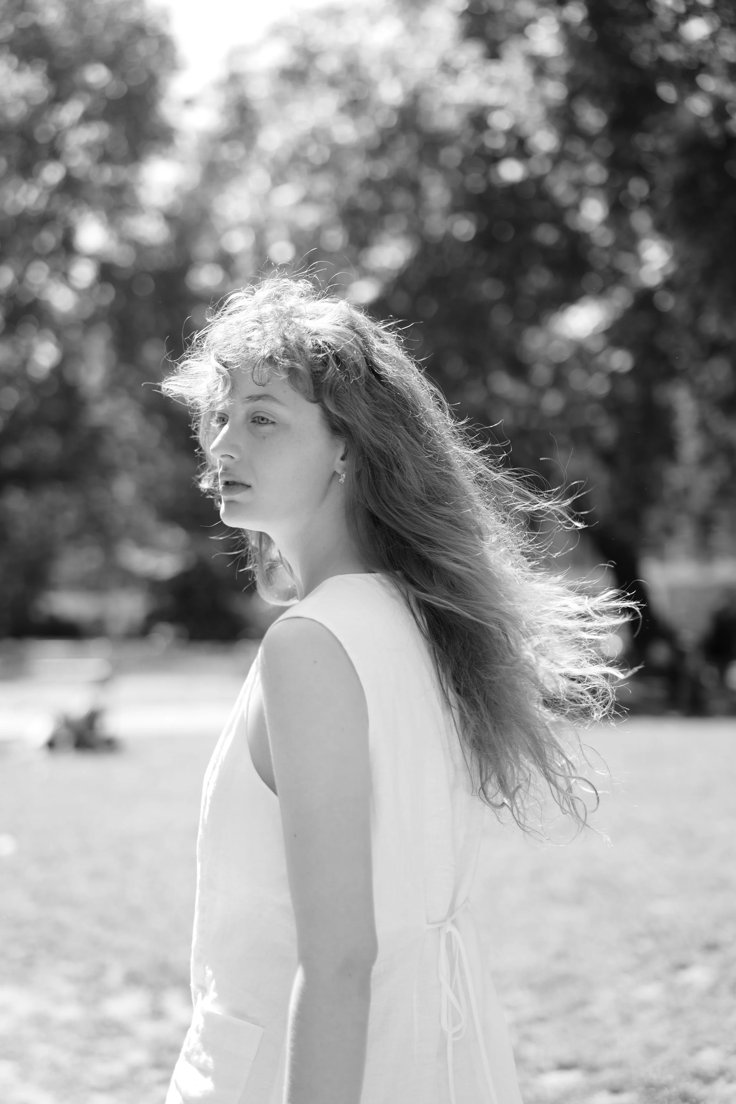 A black and white photo of a young woman with curly hair standing outdoors on a sunny day, with a blurred background of trees and grass.
