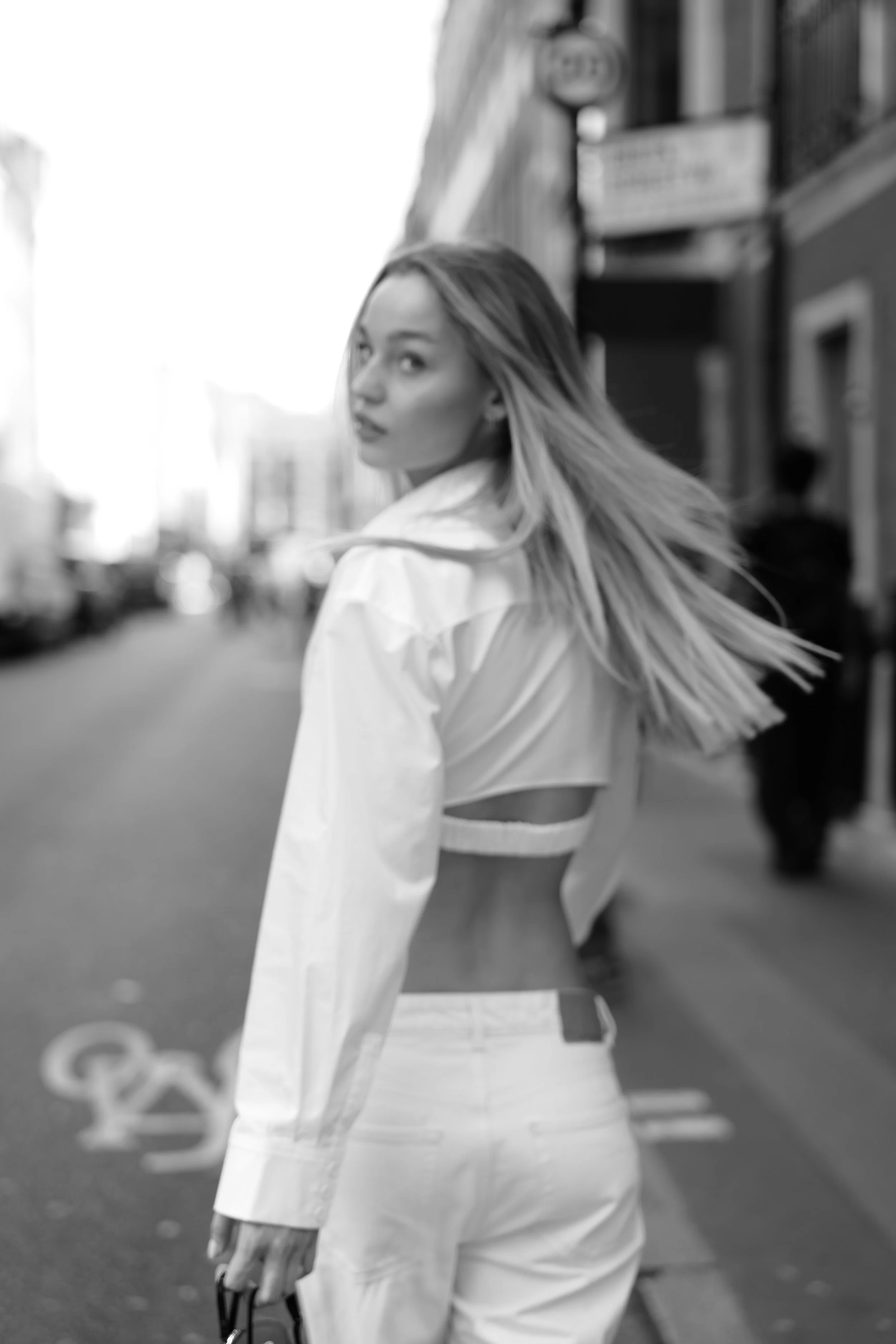 A woman with long hair looking over her shoulder on a city street, wearing a white jacket and holding sunglasses, with buildings and blurred pedestrians in the background.