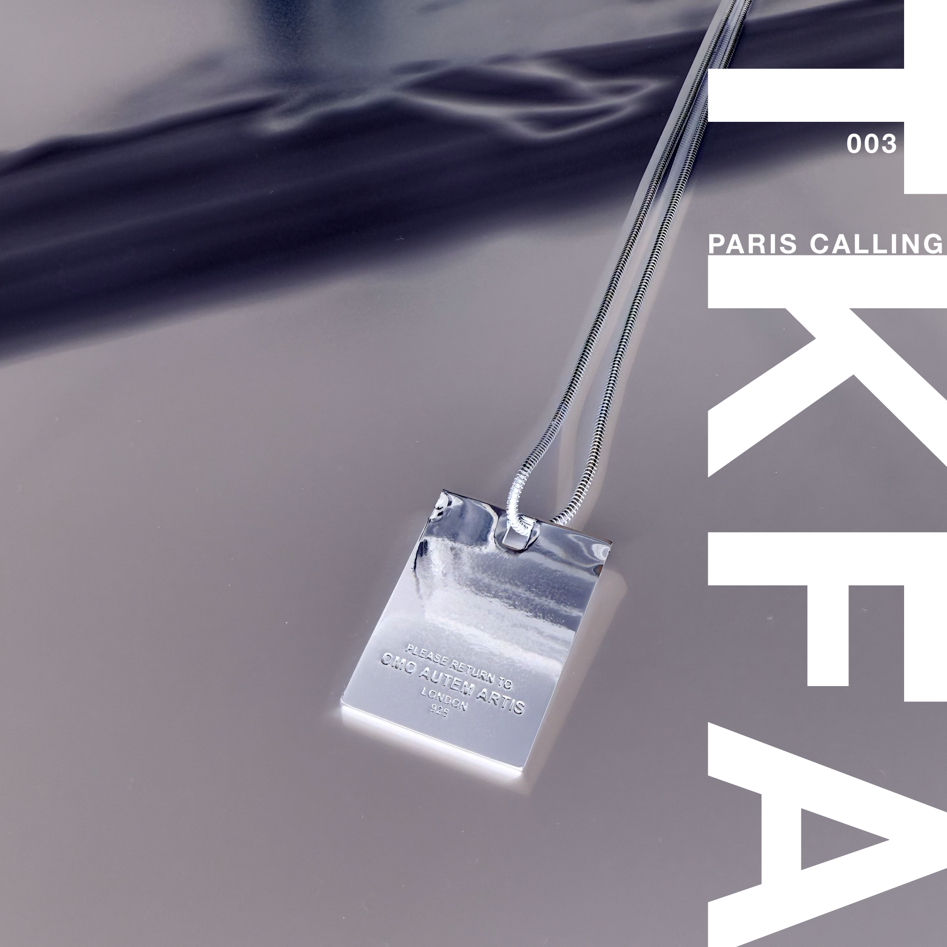 Close-up of a silver necklace with a pendant engraved with the message 'PLEASE RETURN TO OMO AUTEM ARTIS LONDON 925' resting on a smooth, reflective surface.