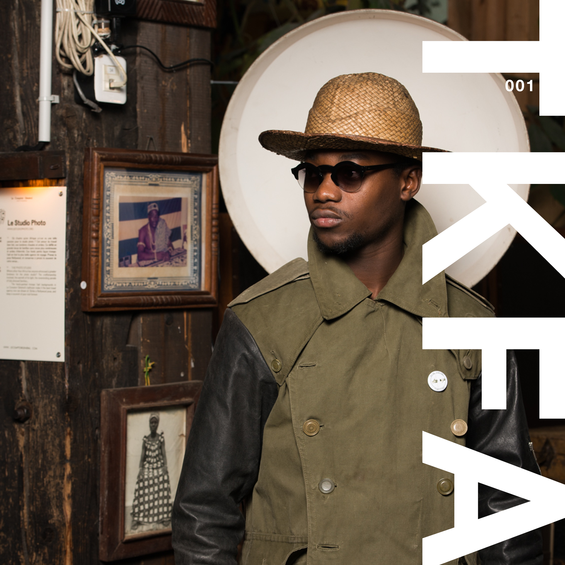 A young man wearing a straw hat, black sunglasses, and a two-tone jacket standing inside a gallery or museum. Behind him is a brown wall with framed photographs and a large, white circular object.
