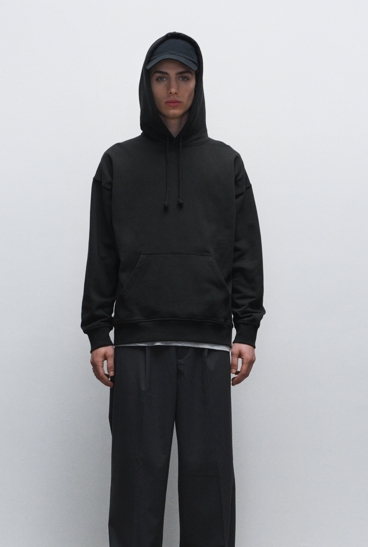 Young man wearing a black hoodie, baseball cap, and loose black pants, standing against a plain white wall.