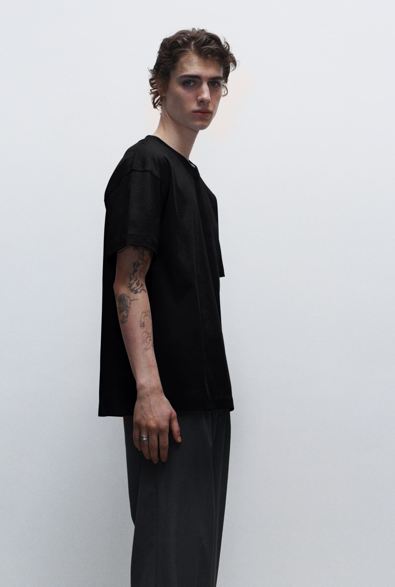 A young man with curly hair and tattoos on his arm, wearing a black t-shirt and dark pants, standing against a plain white background.