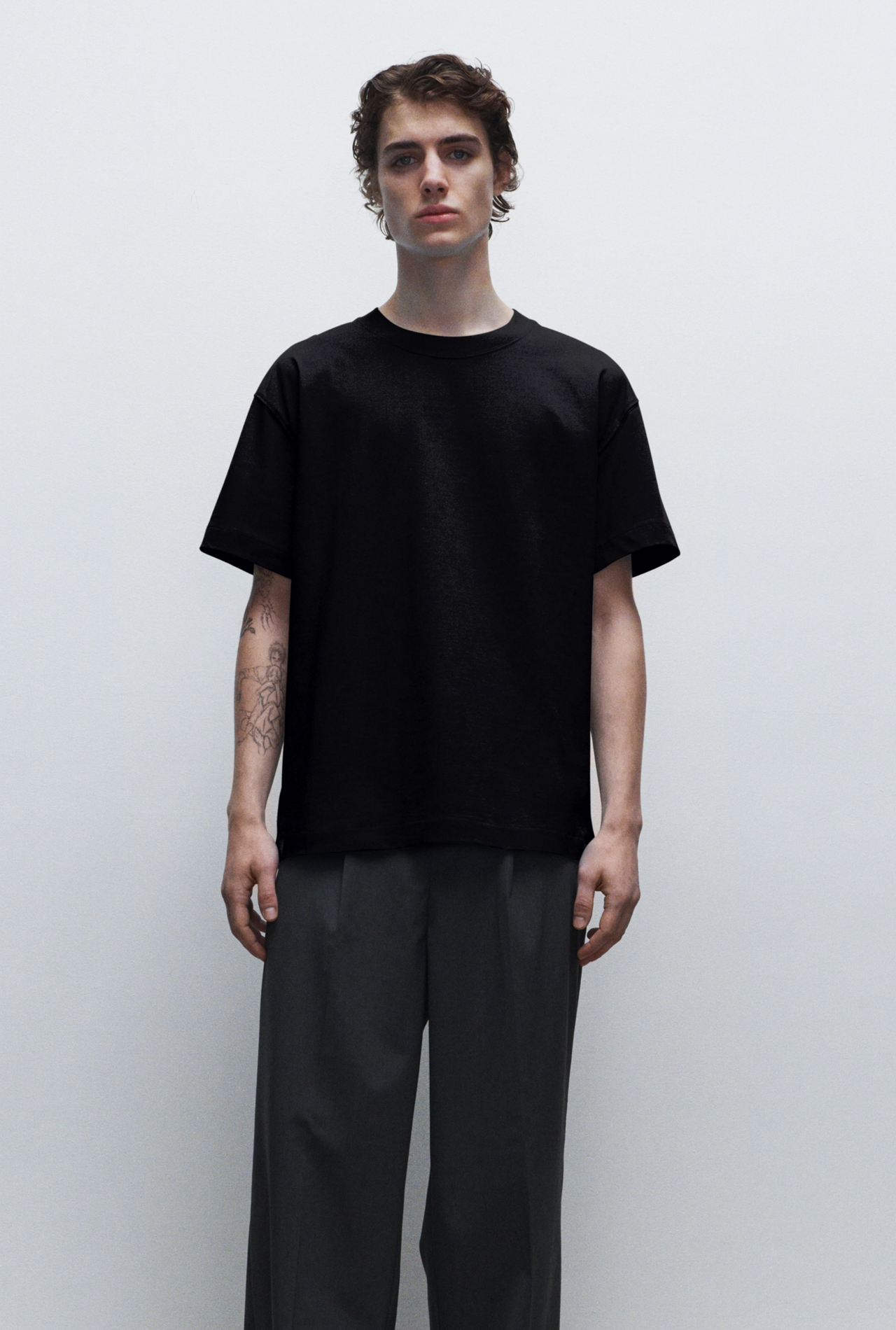 A young person with tattoos on one arm, wearing a black T-shirt and dark gray pants, standing against a plain white background.