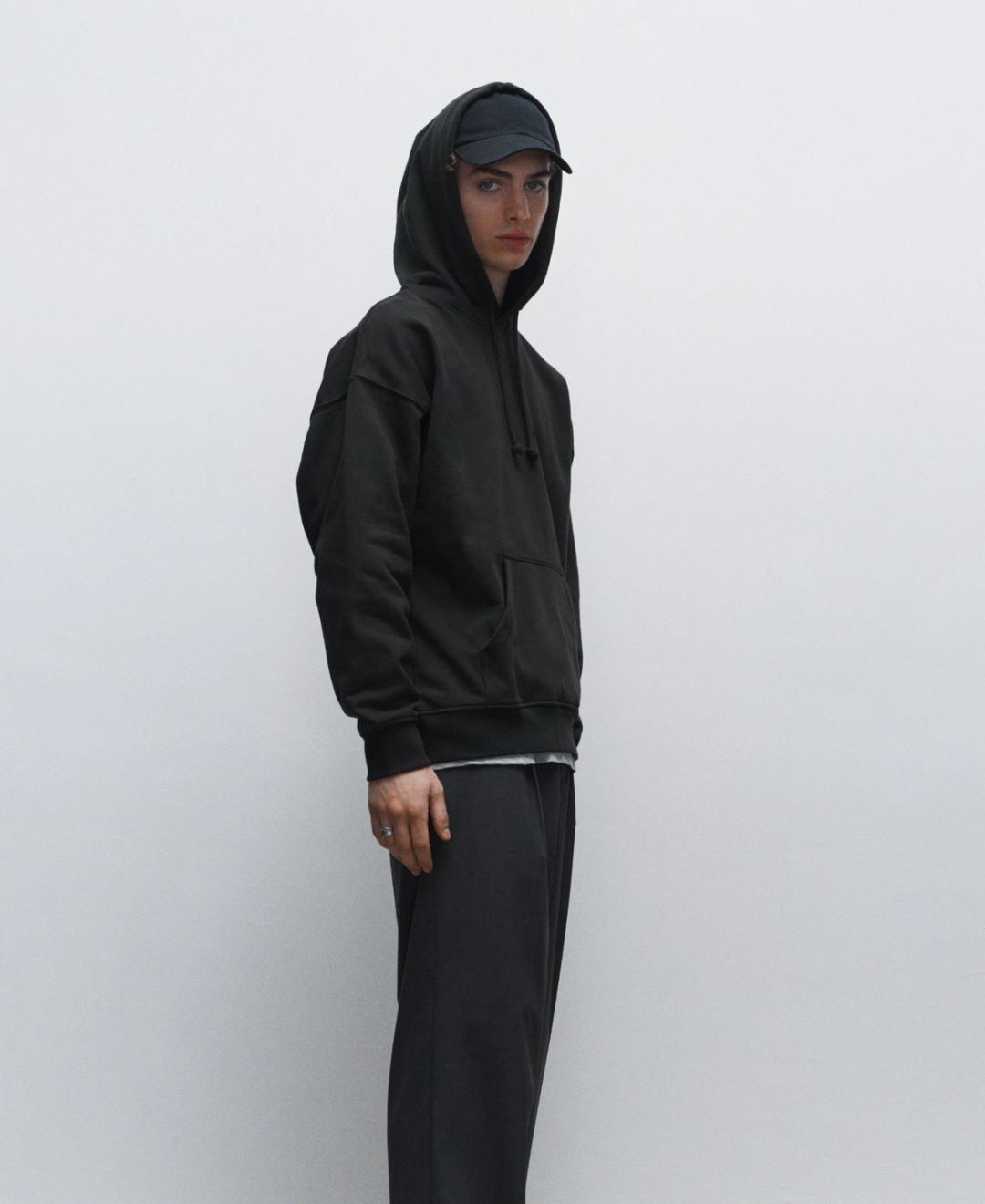 Young man wearing a black hoodie, black cap, and dark pants, standing against a plain, light gray background, looking at the camera.