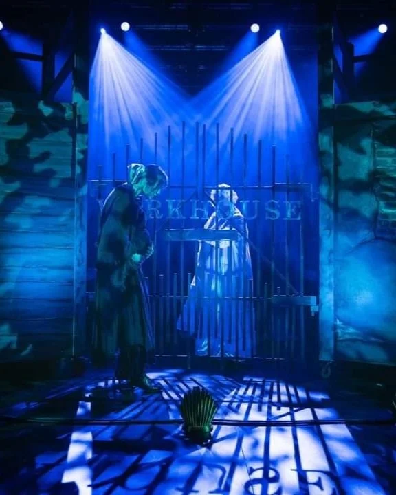 Stage scene with two actors, one inside a cage labeled 'Prisoner,' and the other outside, under blue and purple lighting with shadows cast on the floor and walls.