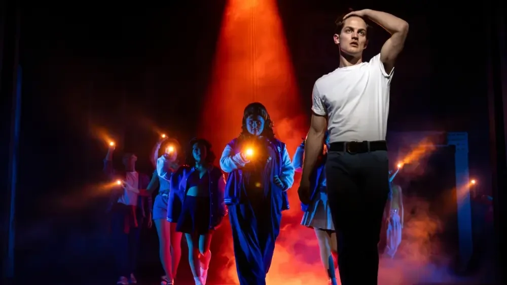 A group of people walking on stage with one man leading, all holding lit torches or flashlights, with dramatic lighting and fog creating a theatrical atmosphere.
