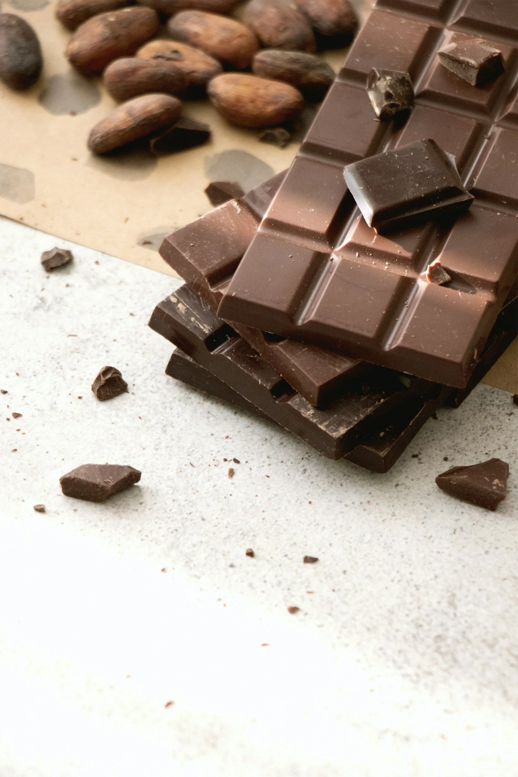 Stacked pieces of dark chocolate with scattered chocolate chips, almonds, and cocoa beans in the background.