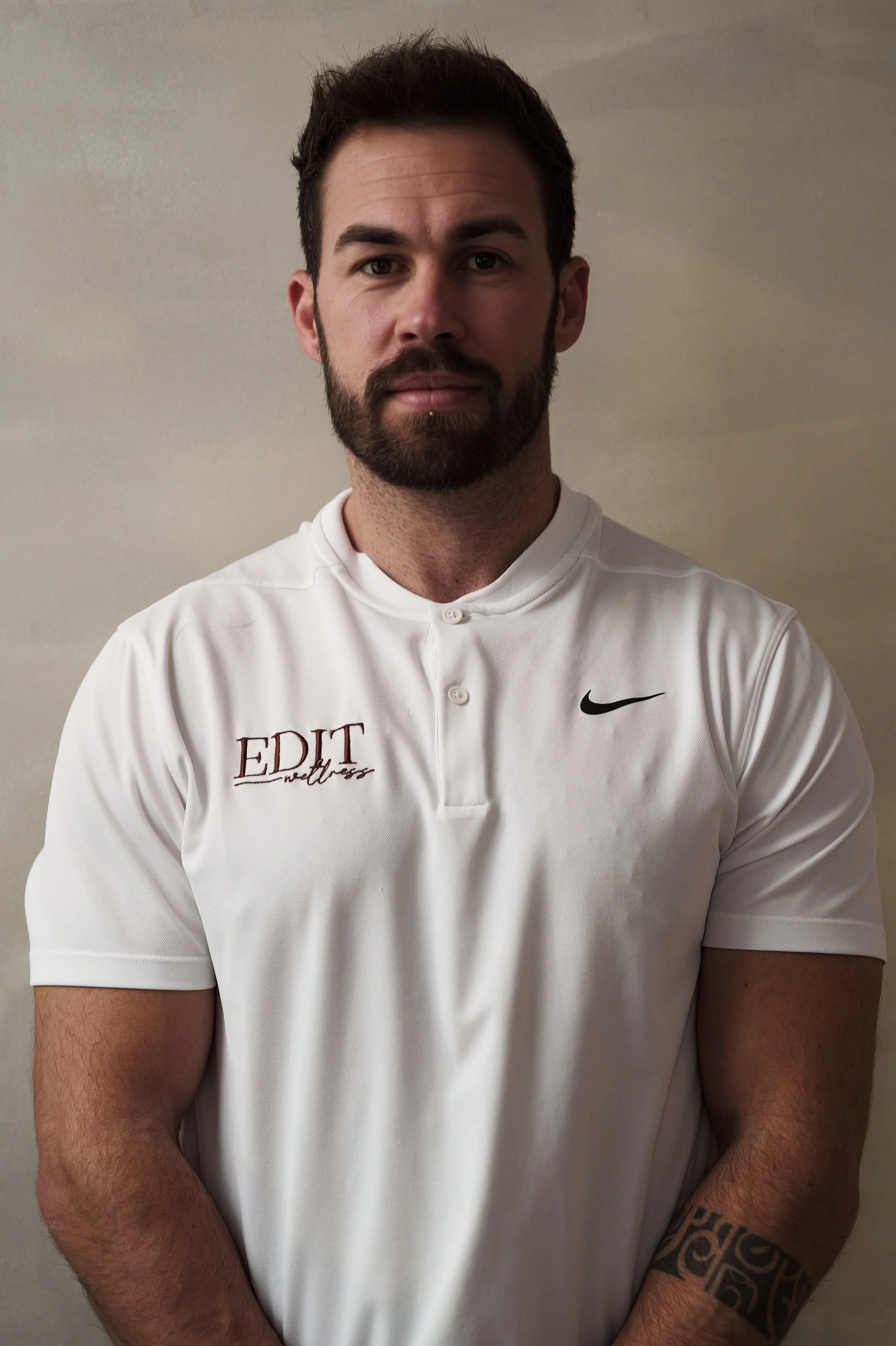 A smiling man with a beard, mustache, and short, curly hair, wearing a white shirt, standing against a plain light-colored background.