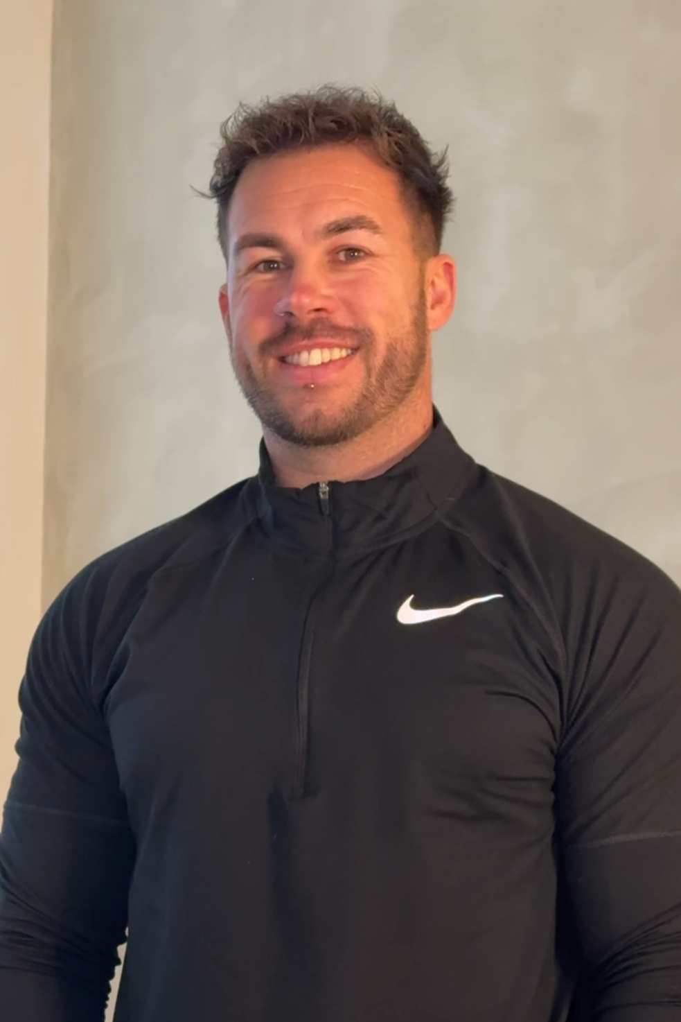 A smiling man with a beard, mustache, and short, curly hair, wearing a white shirt, standing against a plain light-colored background.