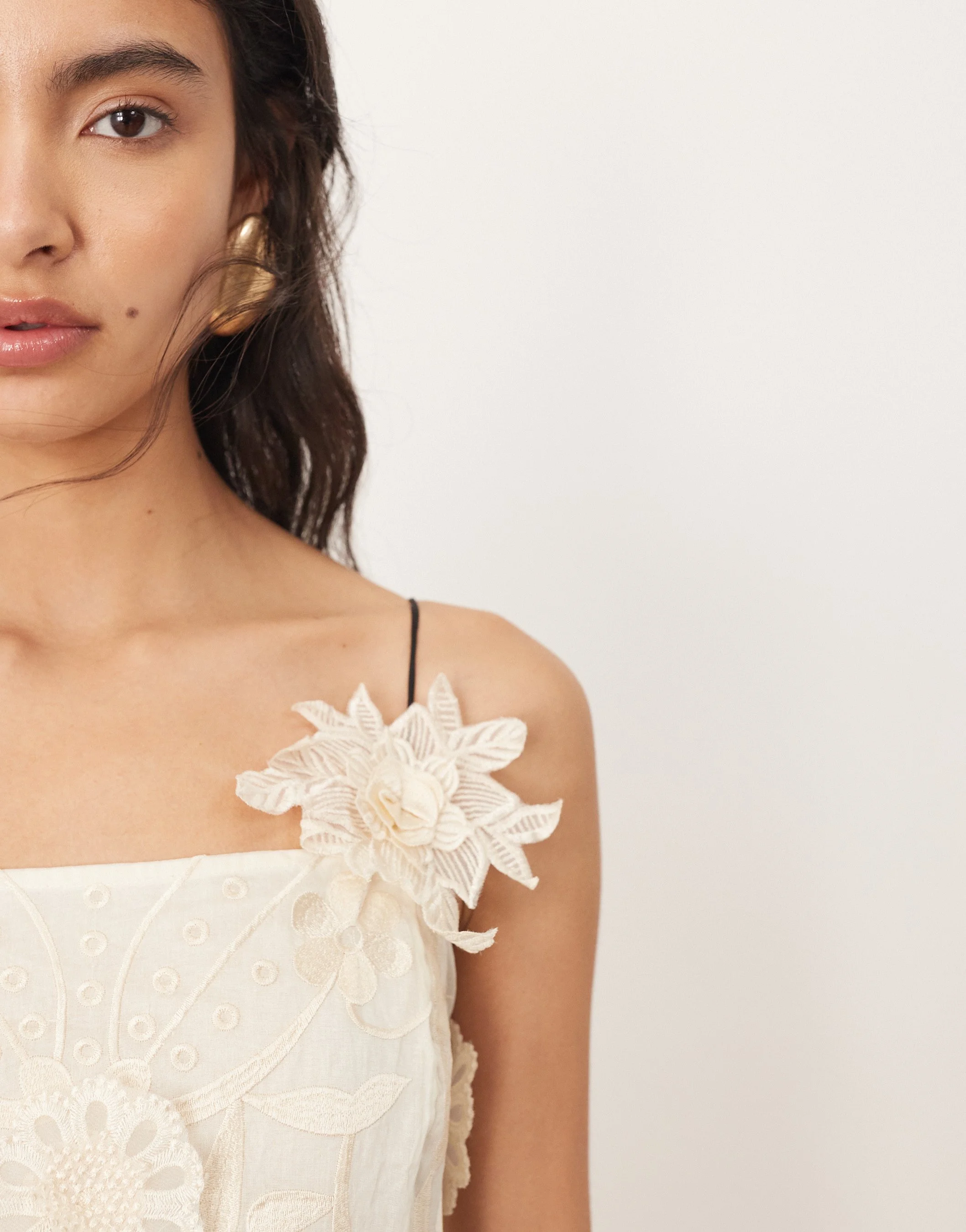 Close-up of a woman's left shoulder, showing a cream-colored dress with embroidered floral designs and a 3D fabric flower on the strap. Part of her face with brown eyes, dark wavy hair, and gold hoop earrings is visible.