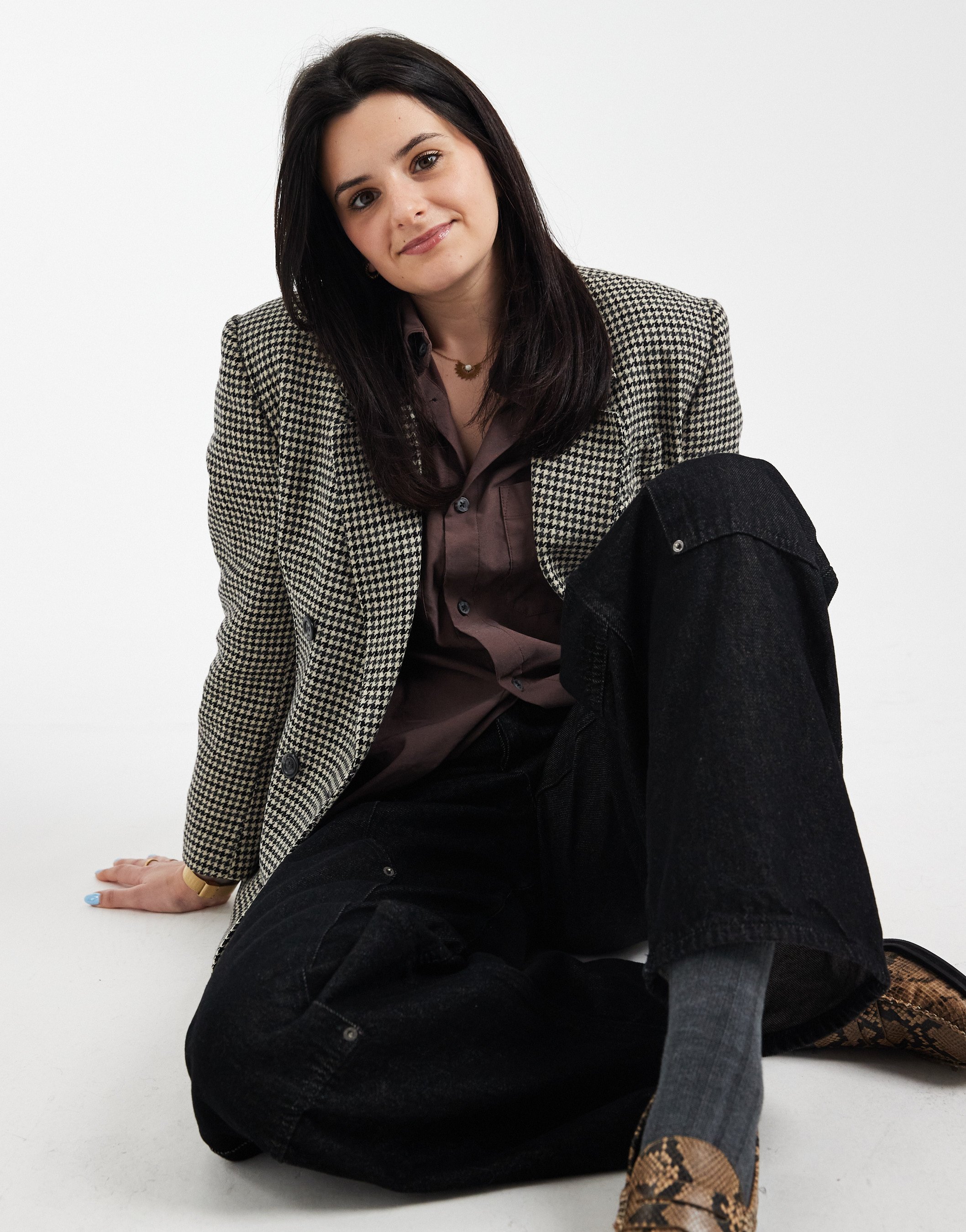 A young woman with dark hair, wearing a checkered blazer over a brown shirt, black jeans, and animal print shoes, sitting on the floor with one leg bent, smiling at the camera against a plain white background.