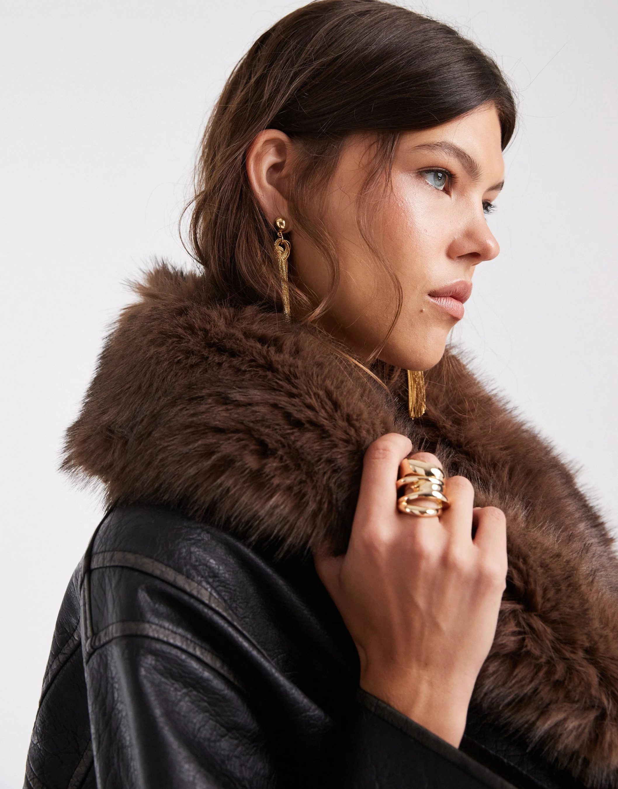 A woman with dark hair, blue eyes, and light skin wearing gold jewelry, a brown fur collar, and a black leather jacket.