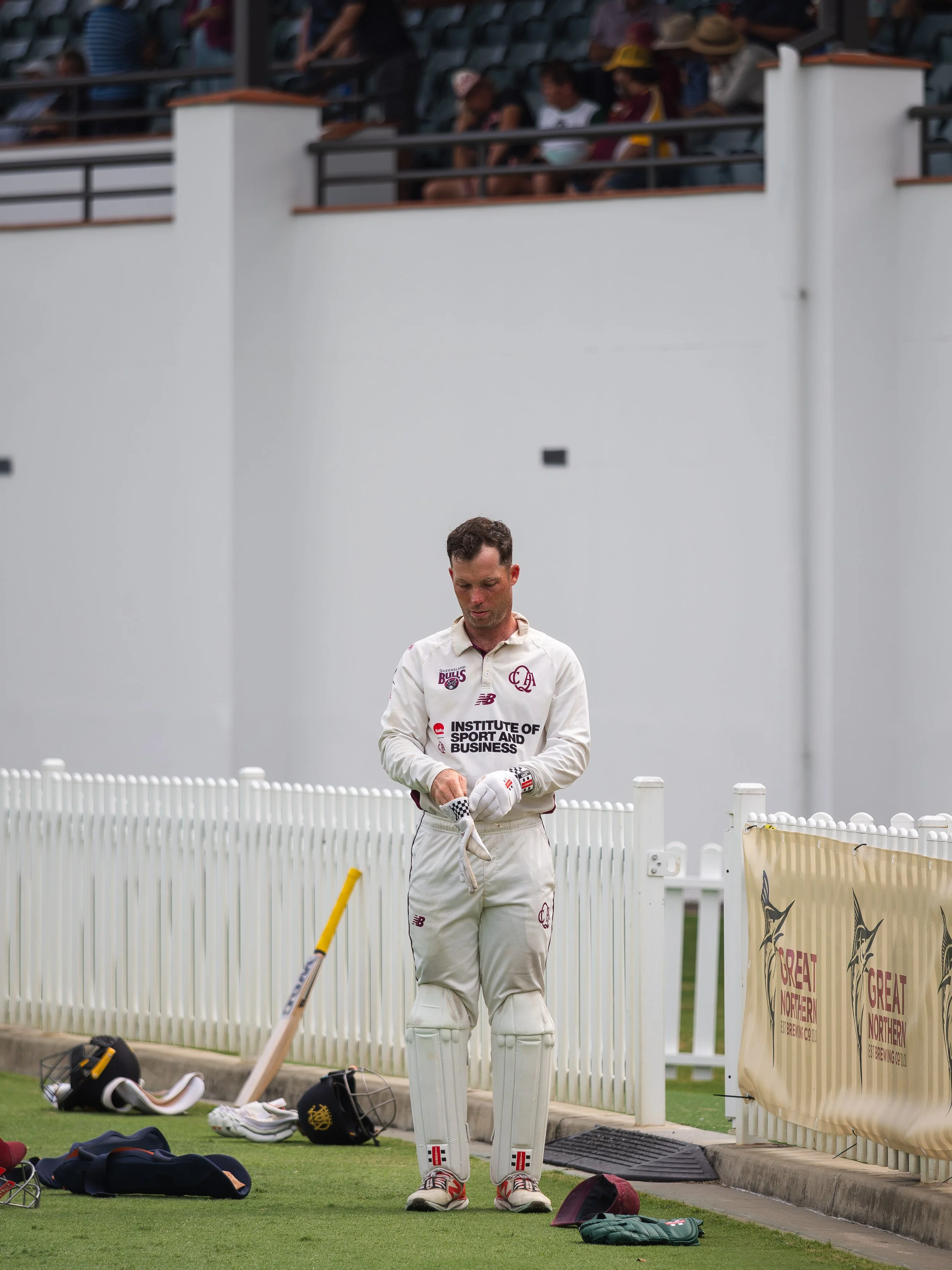 Cricketer standing on field in cricket uniform, preparing for game, with equipment bags and gear on ground, and spectators watching from stands in background.