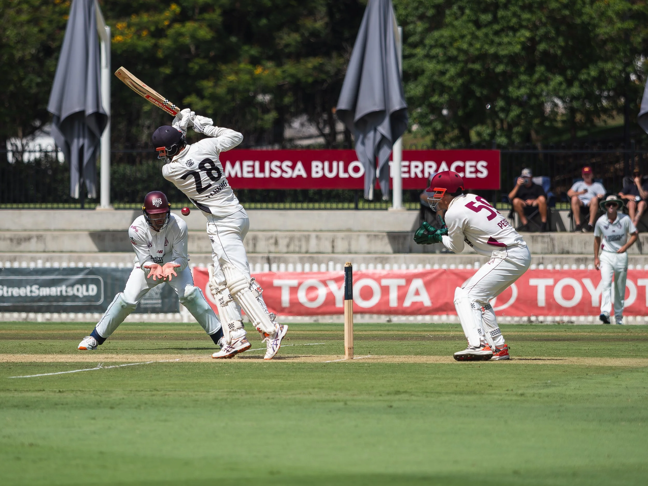 A cricket match in progress with players dressed in white uniforms and helmets on a grassy field. One player is preparing to hit the ball, while others are positioned for fielding. There are spectators in the background watching the game, with some r
