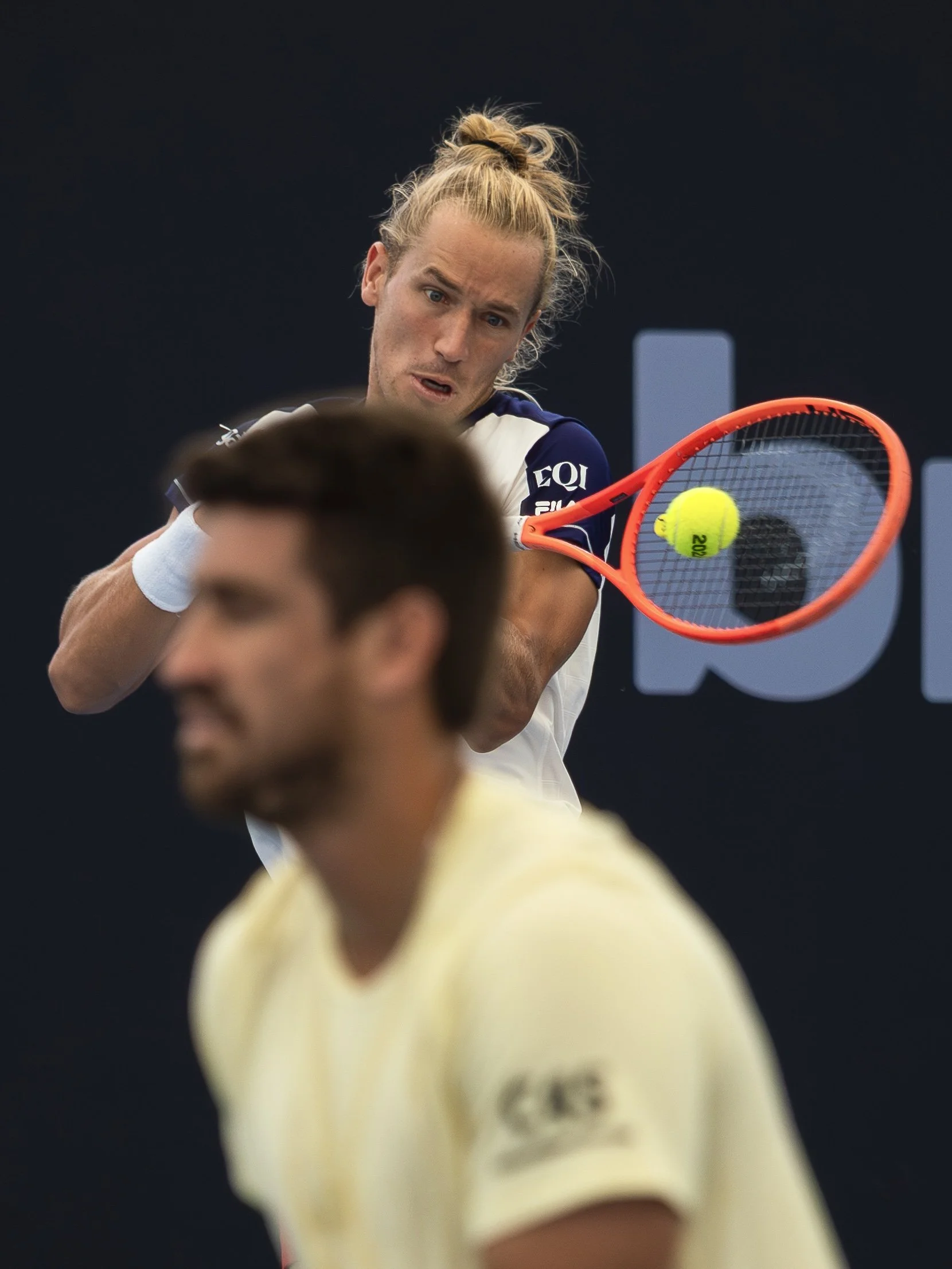 A male tennis player with long blonde hair in a bun is hitting a tennis ball with his racket during a match, wearing a navy white sports outfit against a dark background.
