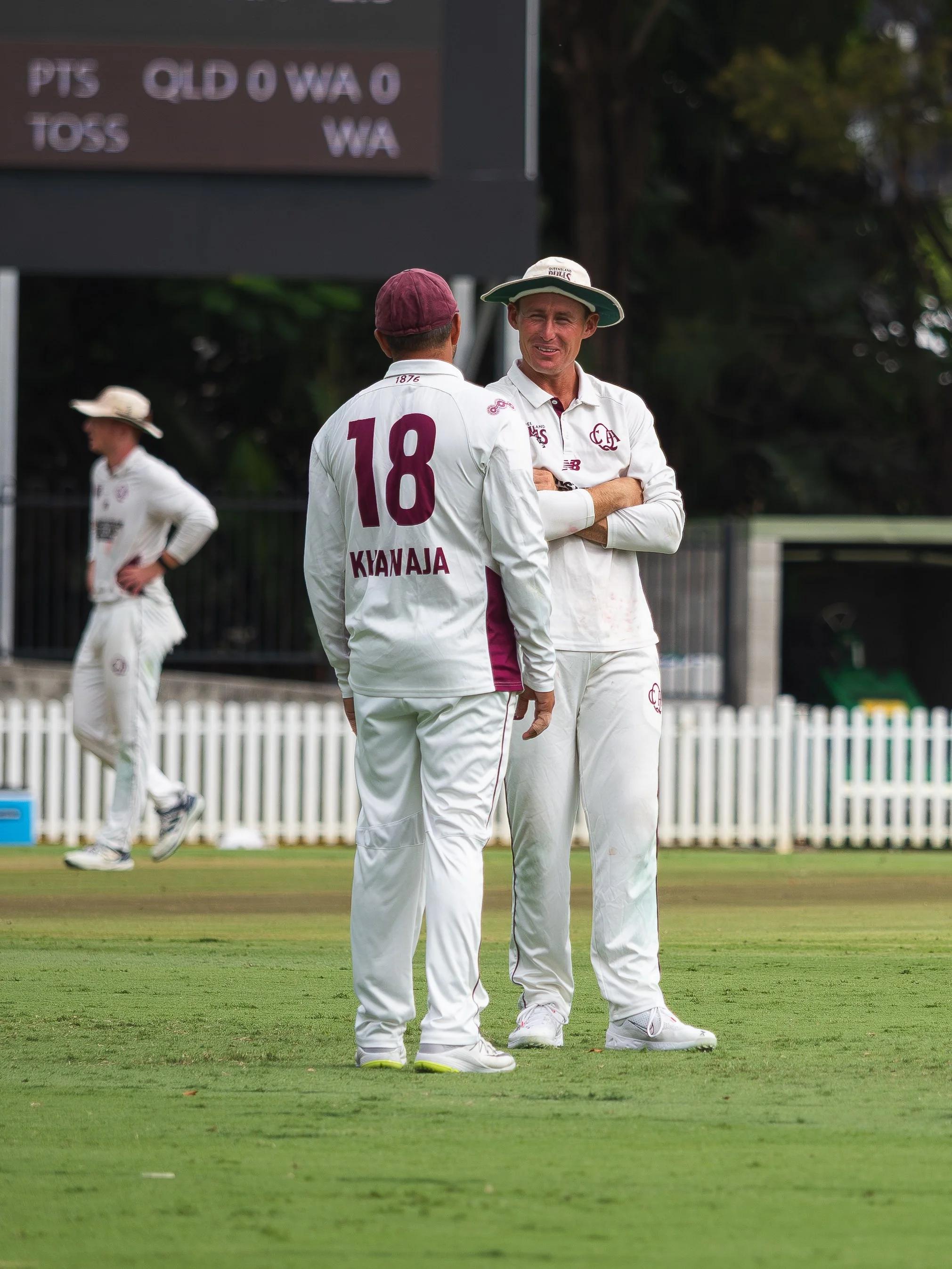 Two cricket players in white uniforms standing on a cricket field, having a conversation. One player, with the number 18 and the name Kwaniaja on the back, is facing another player who is smiling with arms crossed. In the background, a third player i