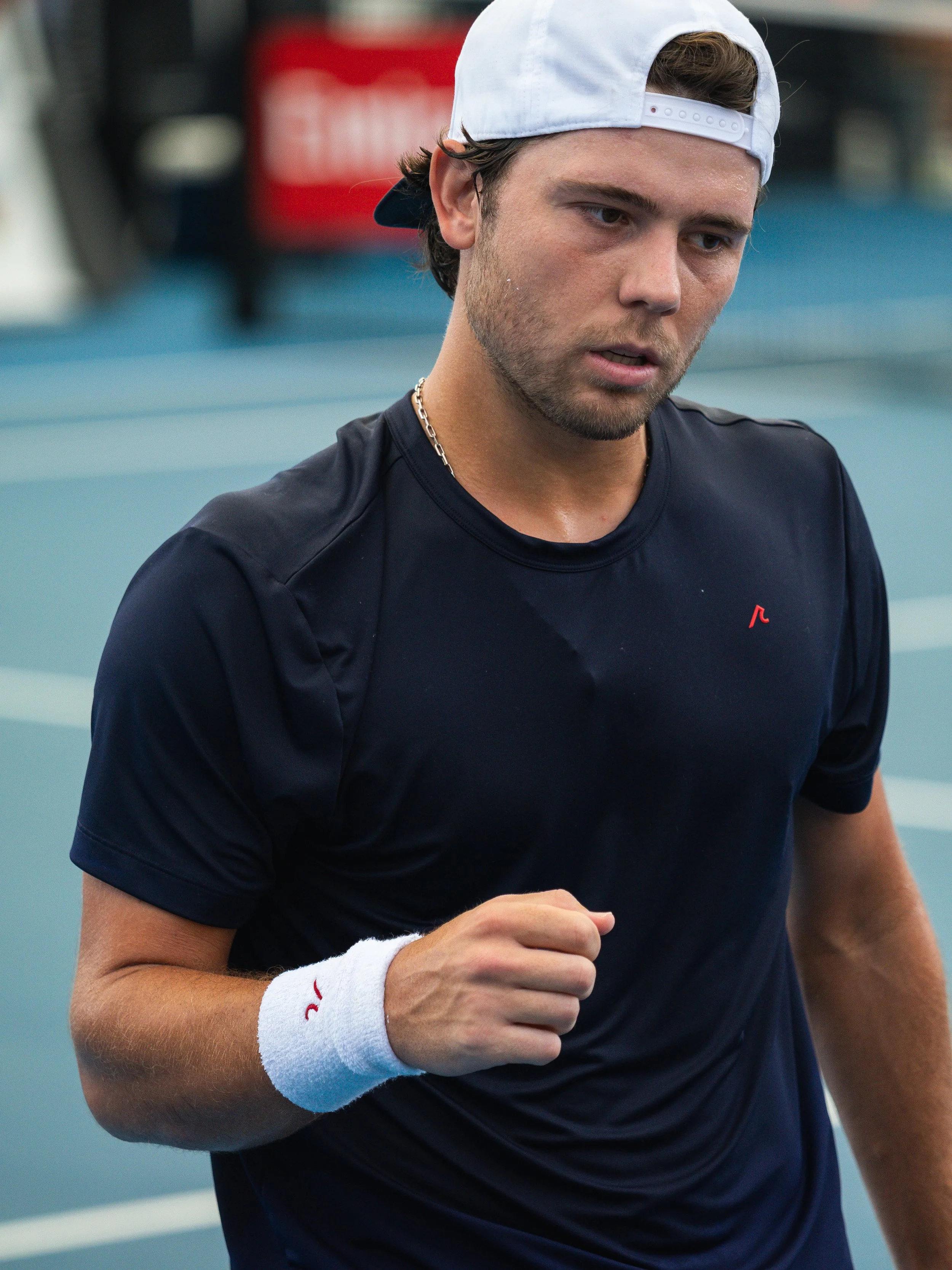 A male tennis player wearing a white cap backward, black sports shirt, and a white sweatband on his wrist, is on a tennis court, appearing focused and in motion.