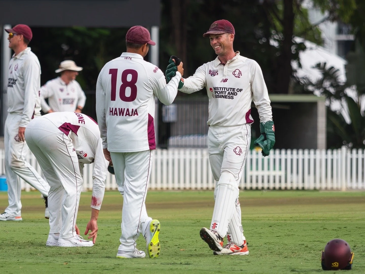 Cricket players on the field, with one player smiling and giving a fist bump to another. The players are dressed in white uniforms with maroon accents, and there is a white picket fence and trees in the background.