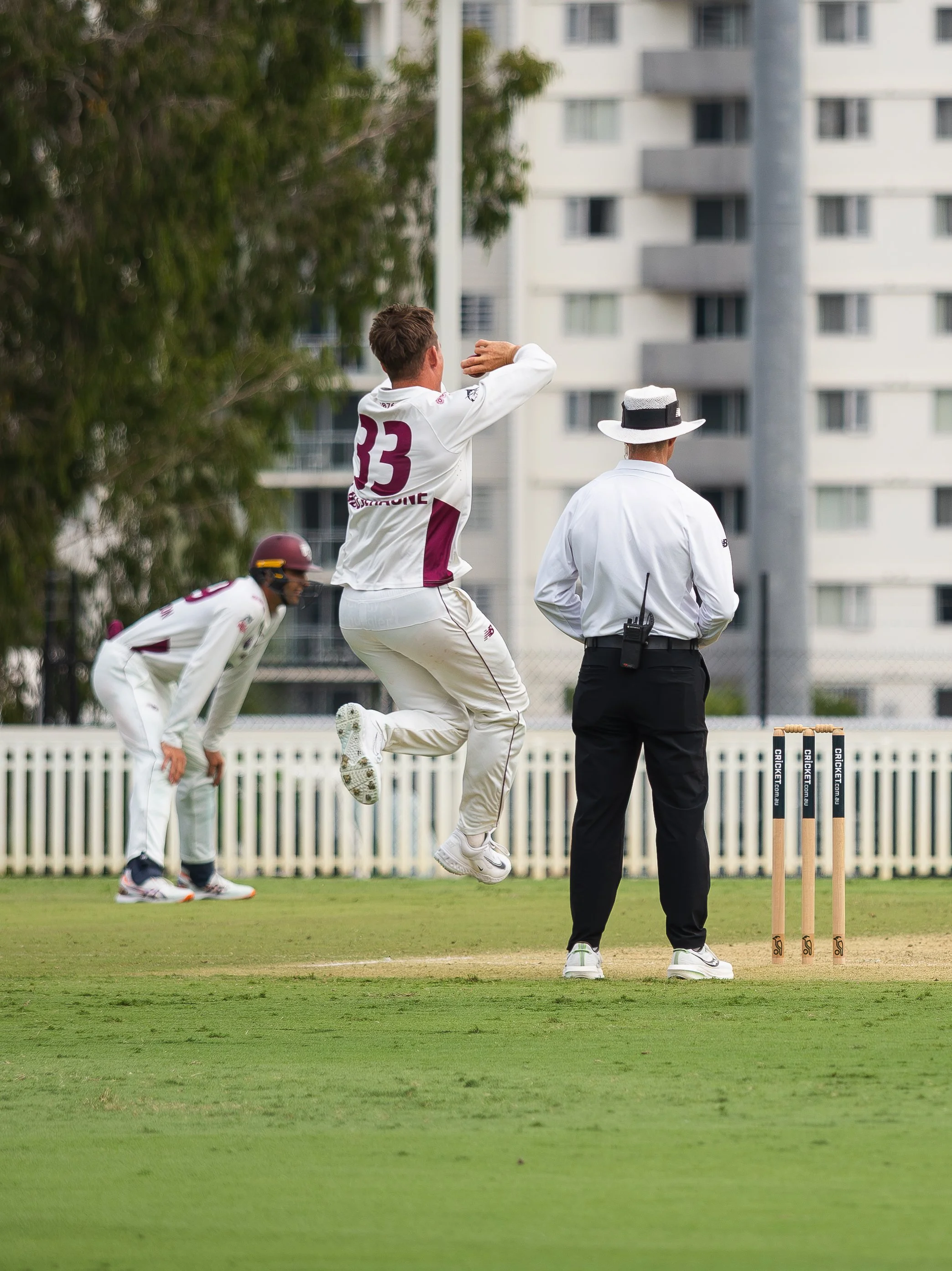 Cricket players on the field with a bowler mid-delivery, an umpire watching, and a background of apartment buildings.