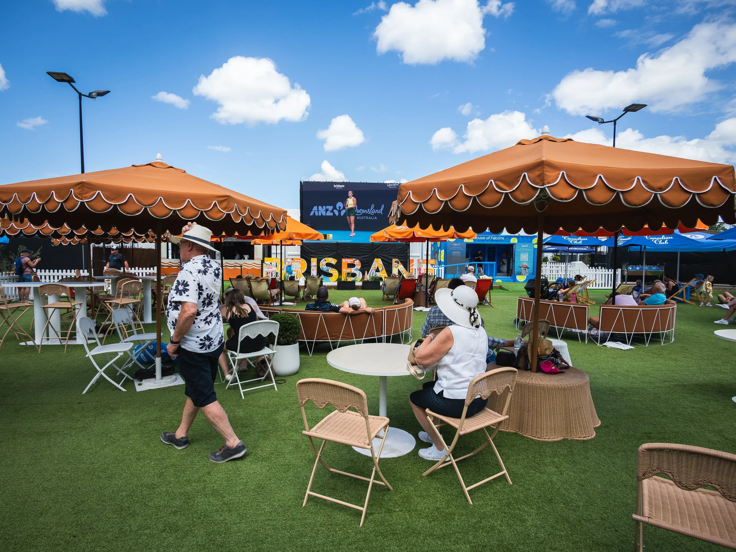 People sitting and walking outdoors at a fair or event under large orange umbrellas with a stage in the background displaying a sign for ANZ and Queensland Australia, with a woman on the screen.