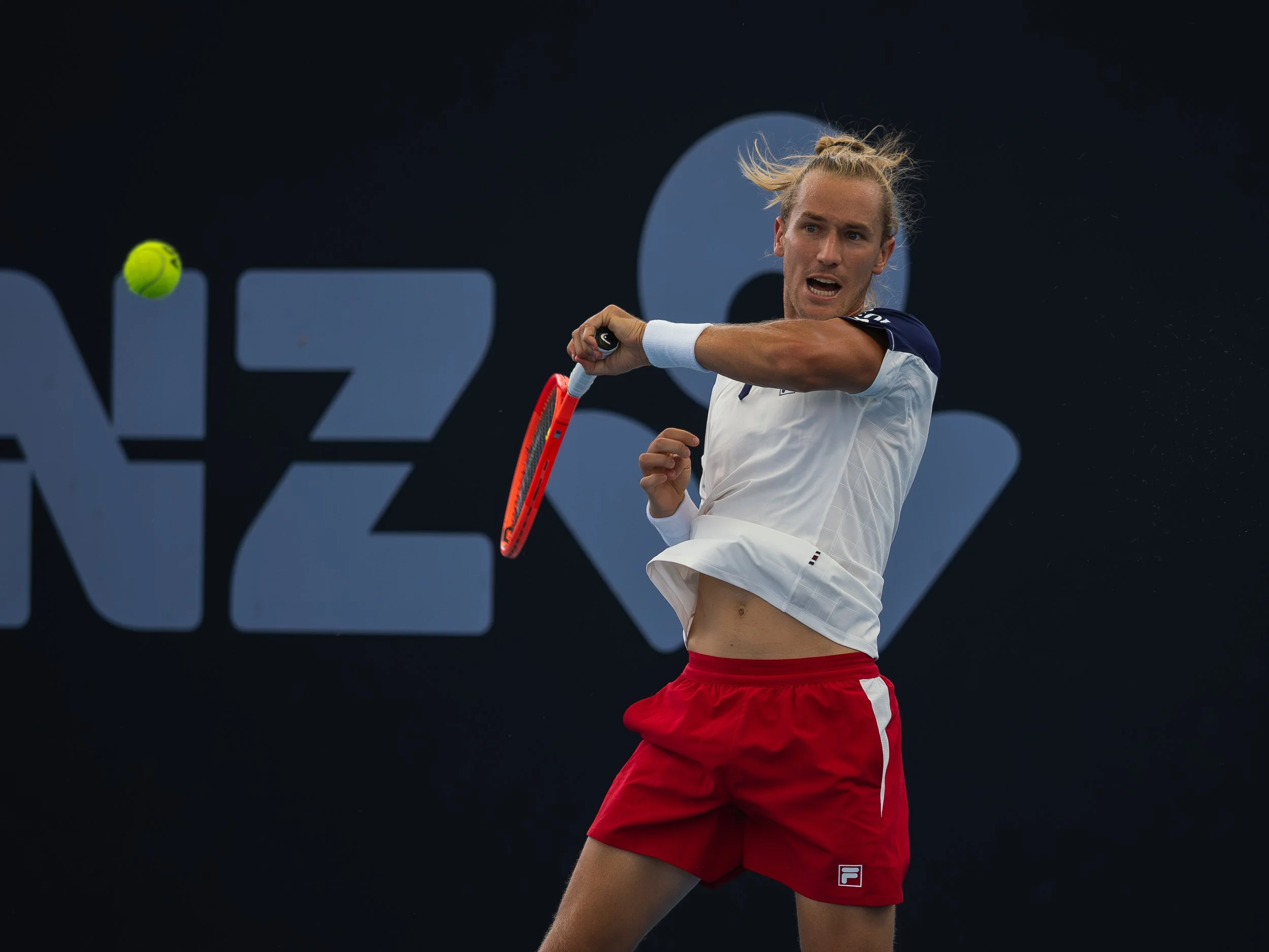 A male tennis player with long blond hair wearing a white and navy tennis shirt and red shorts, hitting a tennis ball with a racquet.