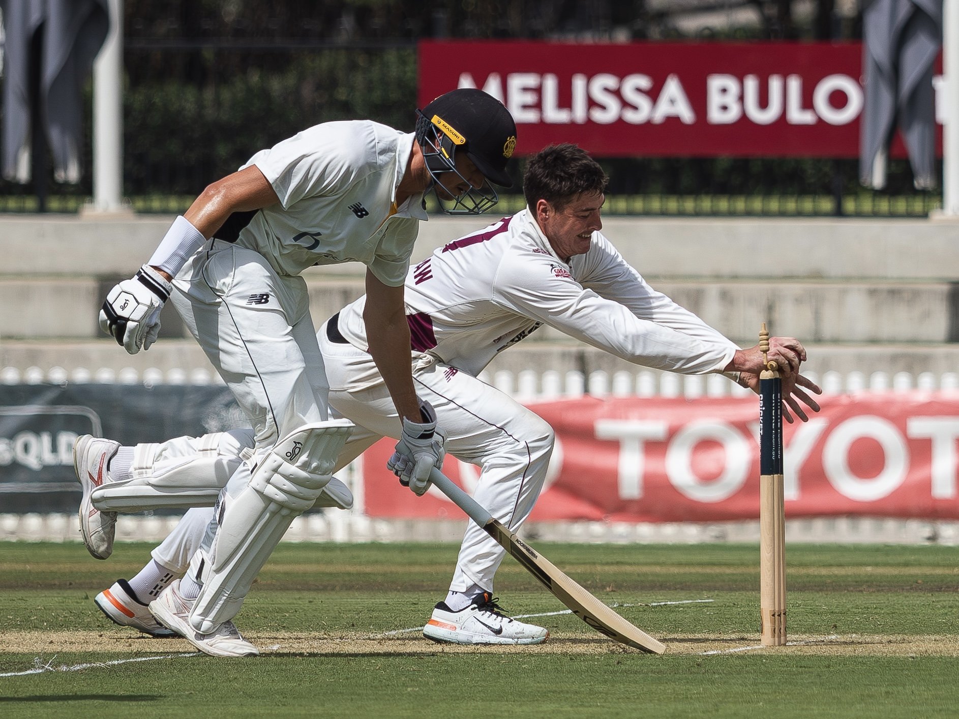 Two cricket players in white uniforms and helmets chase the cricket ball on a grassy field. One player is reaching for the ball while the other is running behind, both focused and in motion.