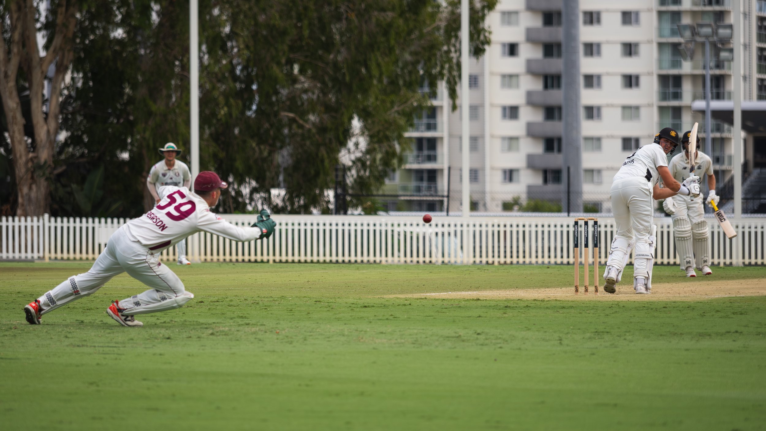 Cricket players in action on the field with two players near the stumps and one player reaching to catch the ball, while other players observe in the background, surrounded by green grass, trees, and apartment buildings.