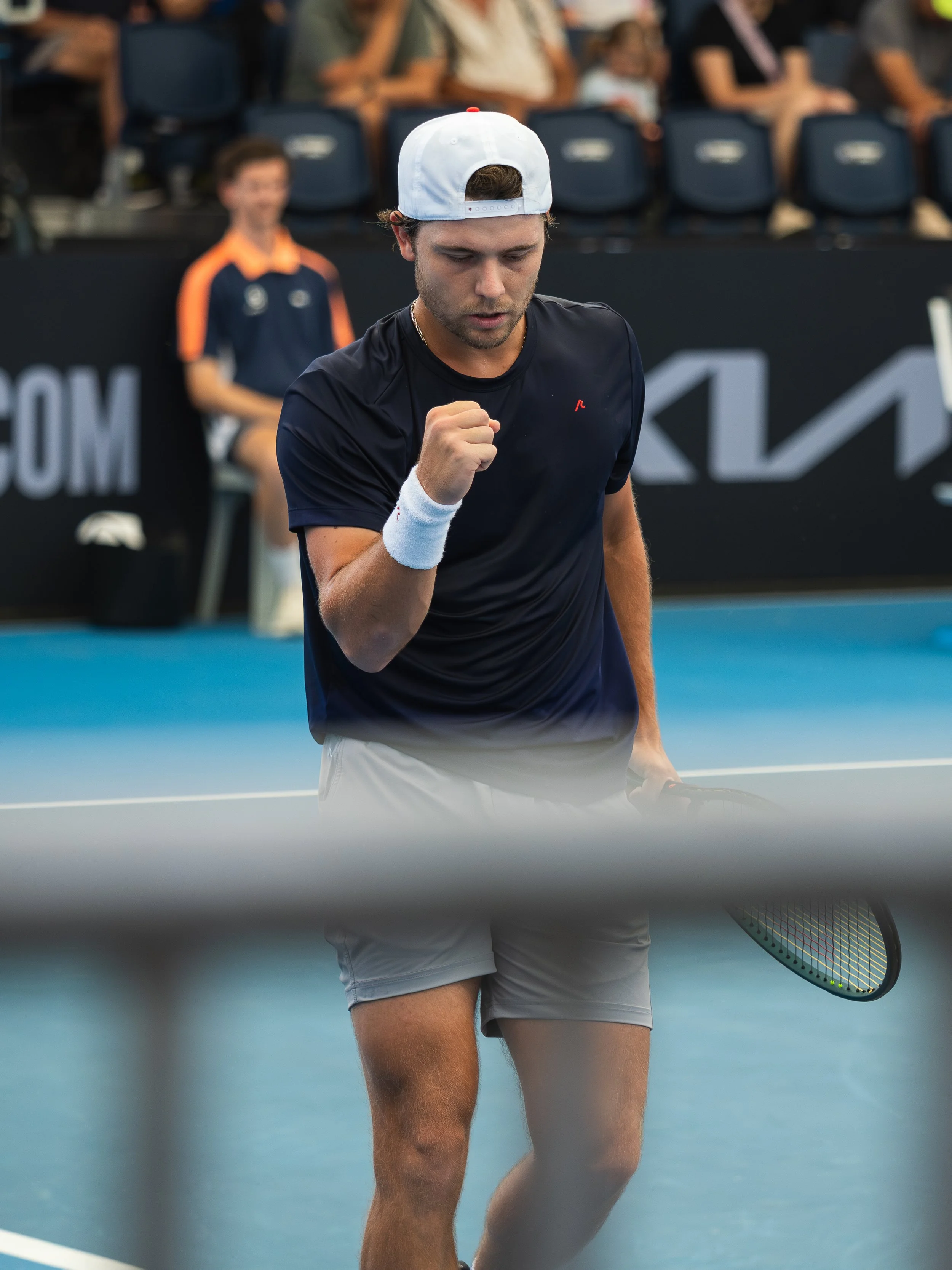 A male tennis player wearing a white cap, black shirt, and light gray shorts, holding a tennis racket, looks down with a clenched fist during a match on a blue tennis court.