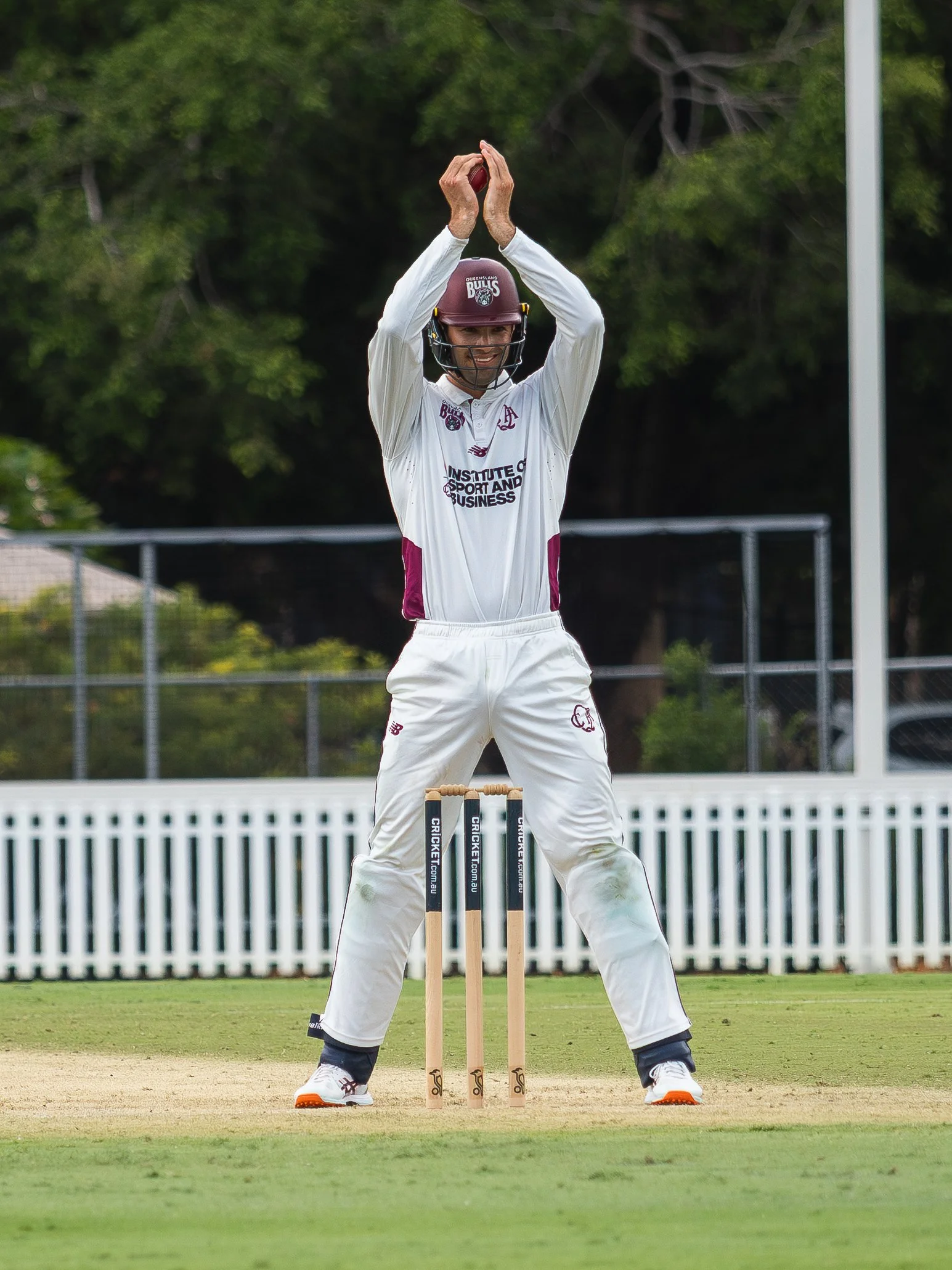 A cricket player in white uniform with maroon helmet preparing to bat on a cricket field, standing in front of the stumps with an overcast sky and greenery in the background.
