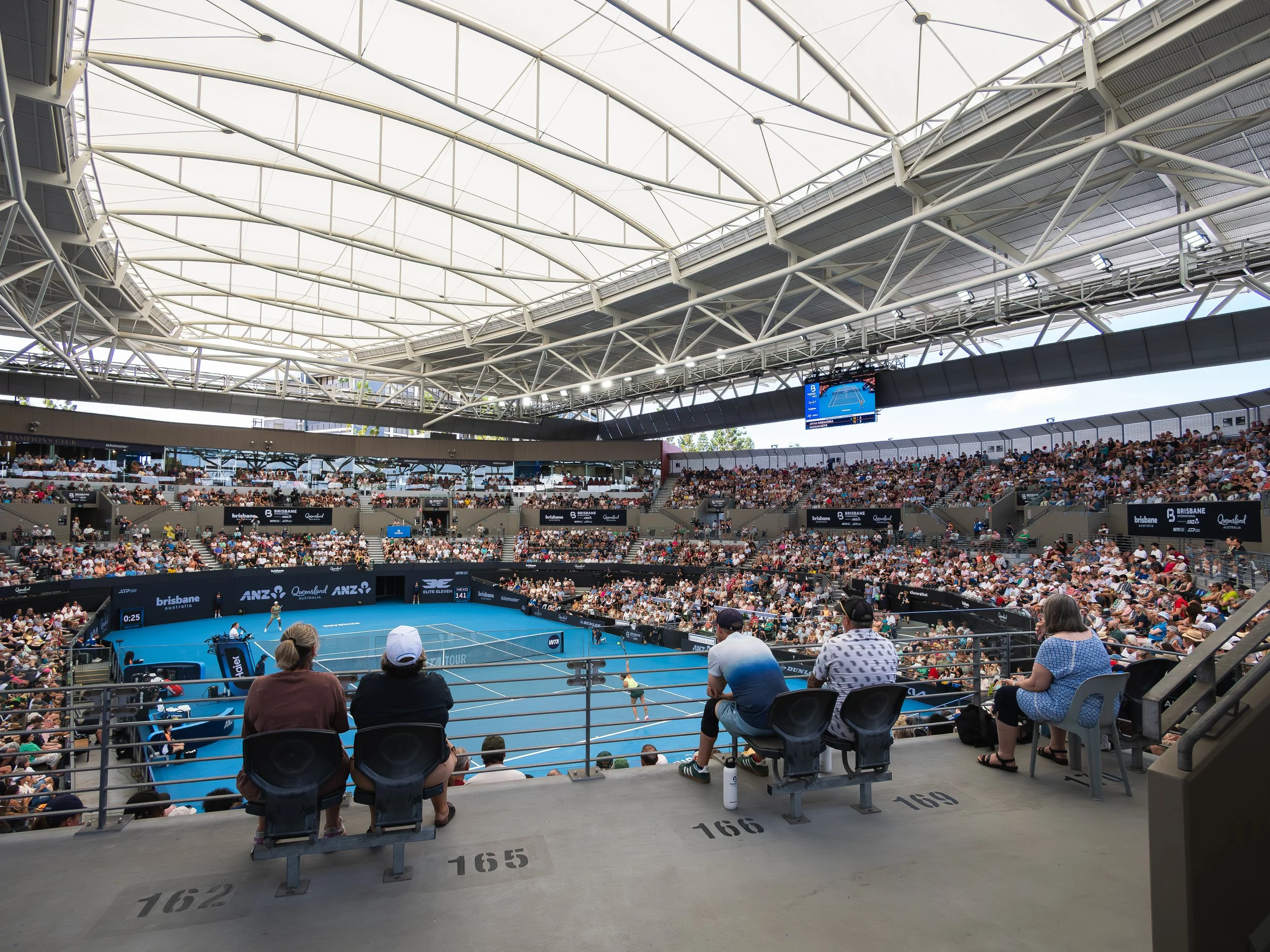 Indoor tennis stadium filled with spectators watching a tennis match on a blue court. The stadium has a modern roof structure with white beams and large lighting fixtures. Some spectators are seated in the foreground, with numbers and sections labele