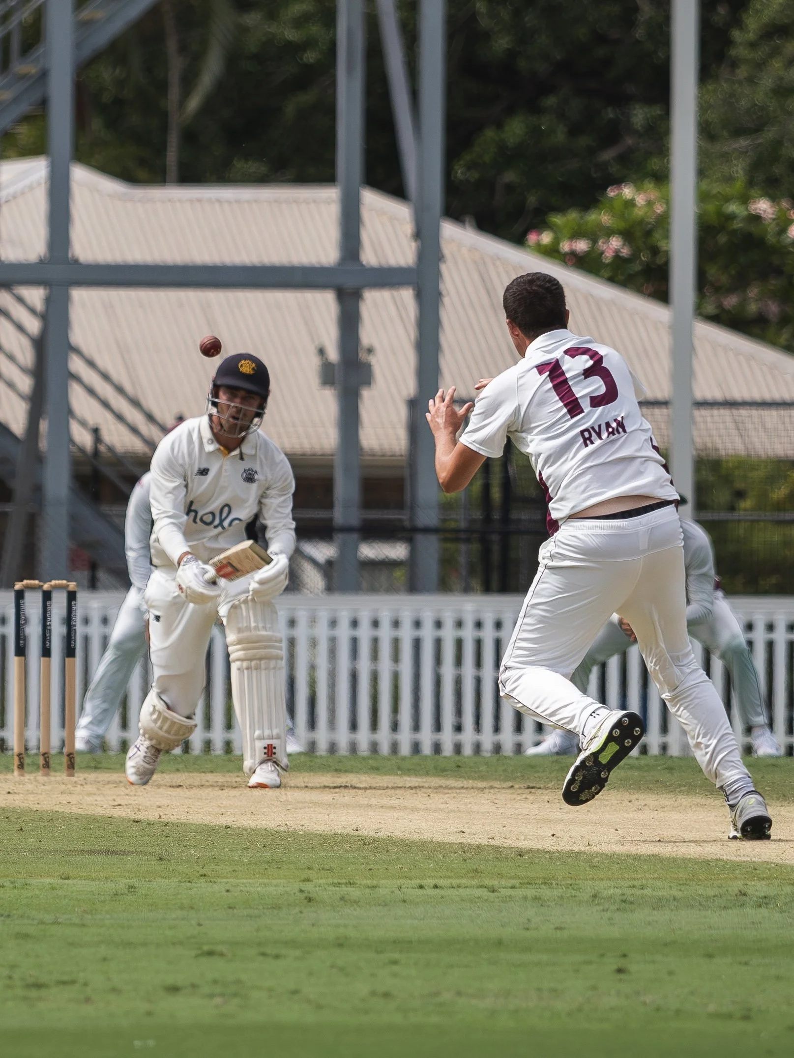 Cricket players in action on the field, one is batting and the other is wicket-keeping, with wickets and a fence in the background.