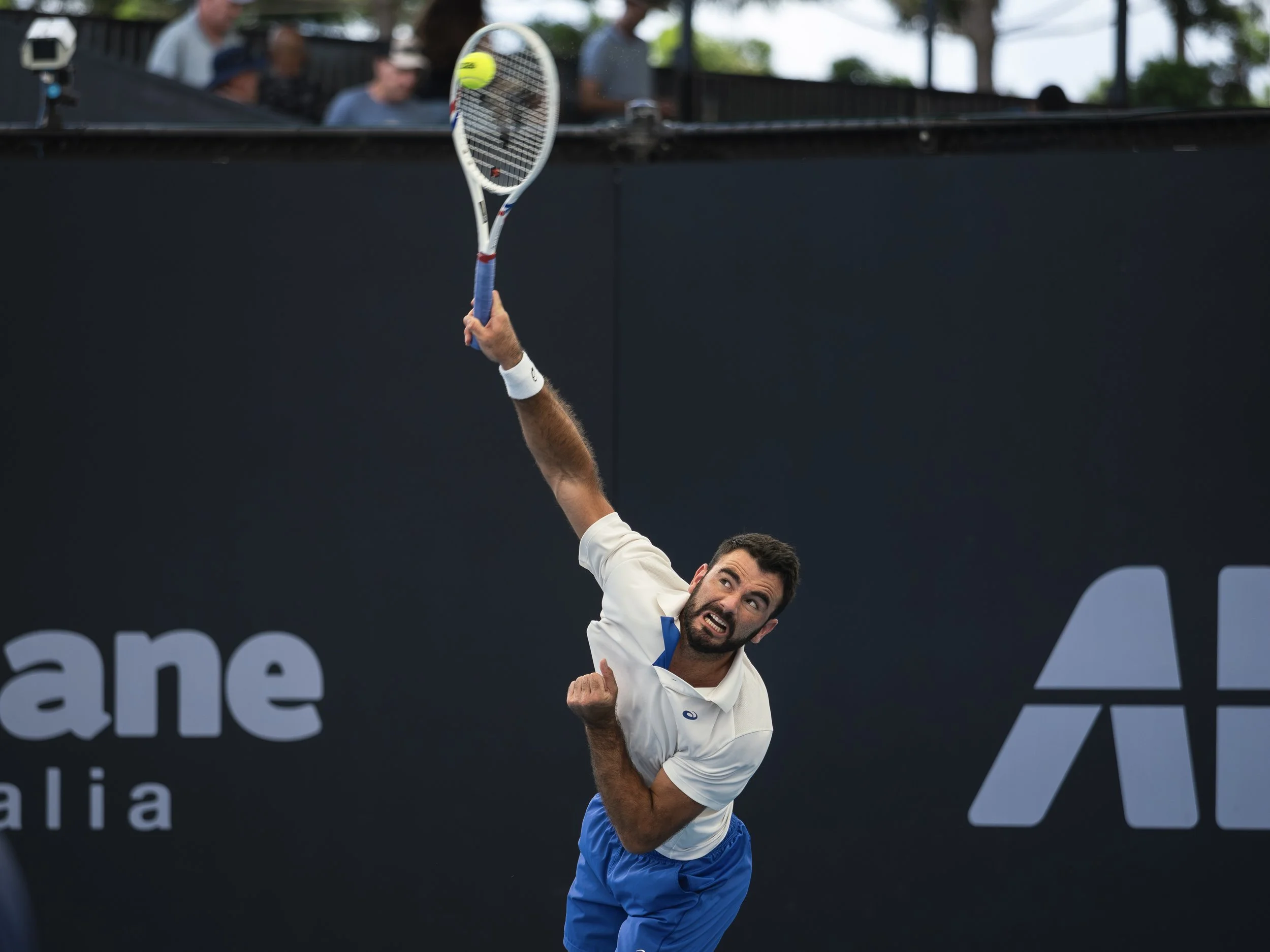 A male tennis player in a white shirt and blue shorts is swinging his tennis racket to hit a tennis ball during a match. The background shows a black wall and some seated spectators.