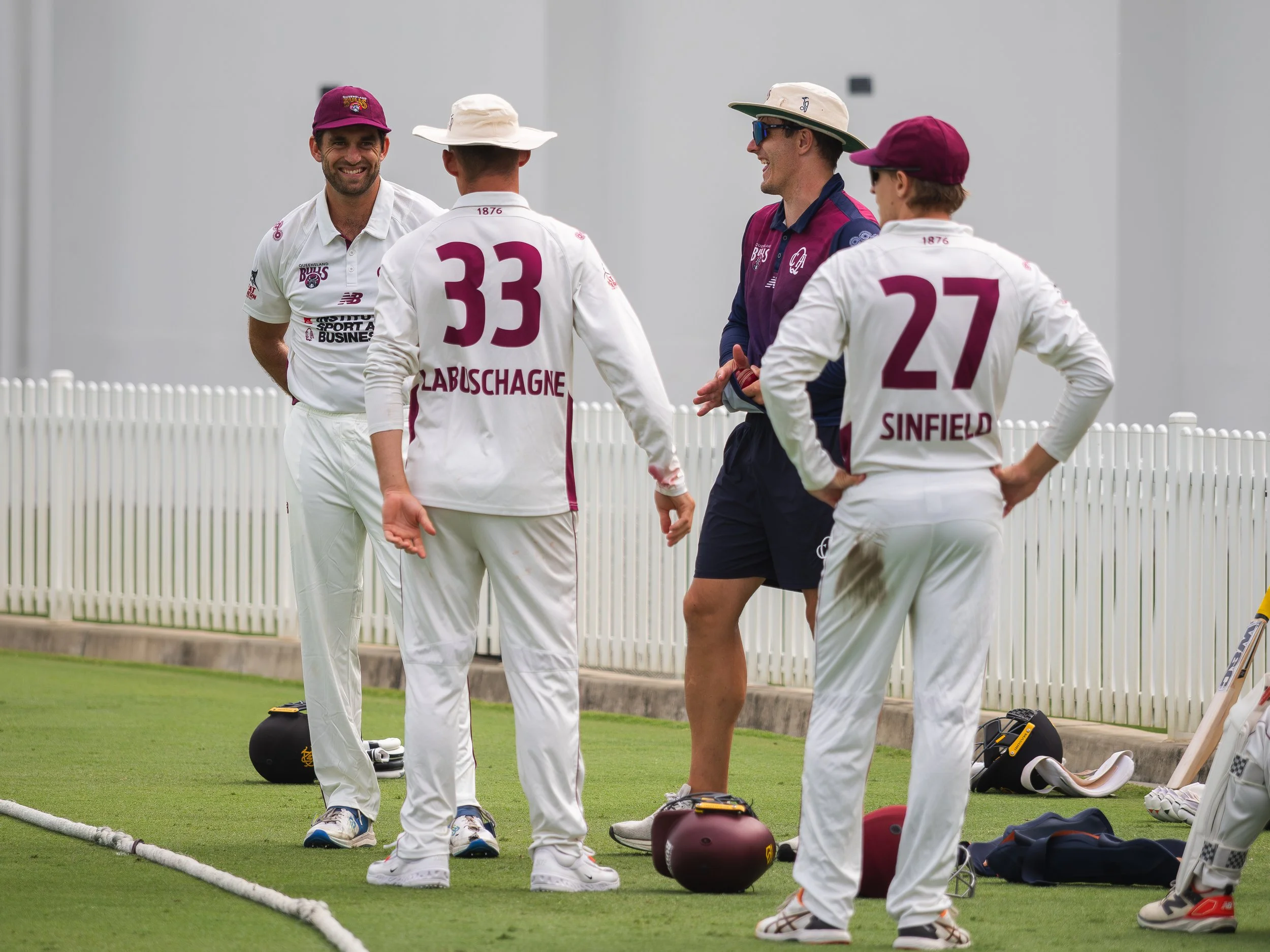 Four men dressed in cricket uniforms standing and talking on a cricket field, with equipment and a white fence in the background.