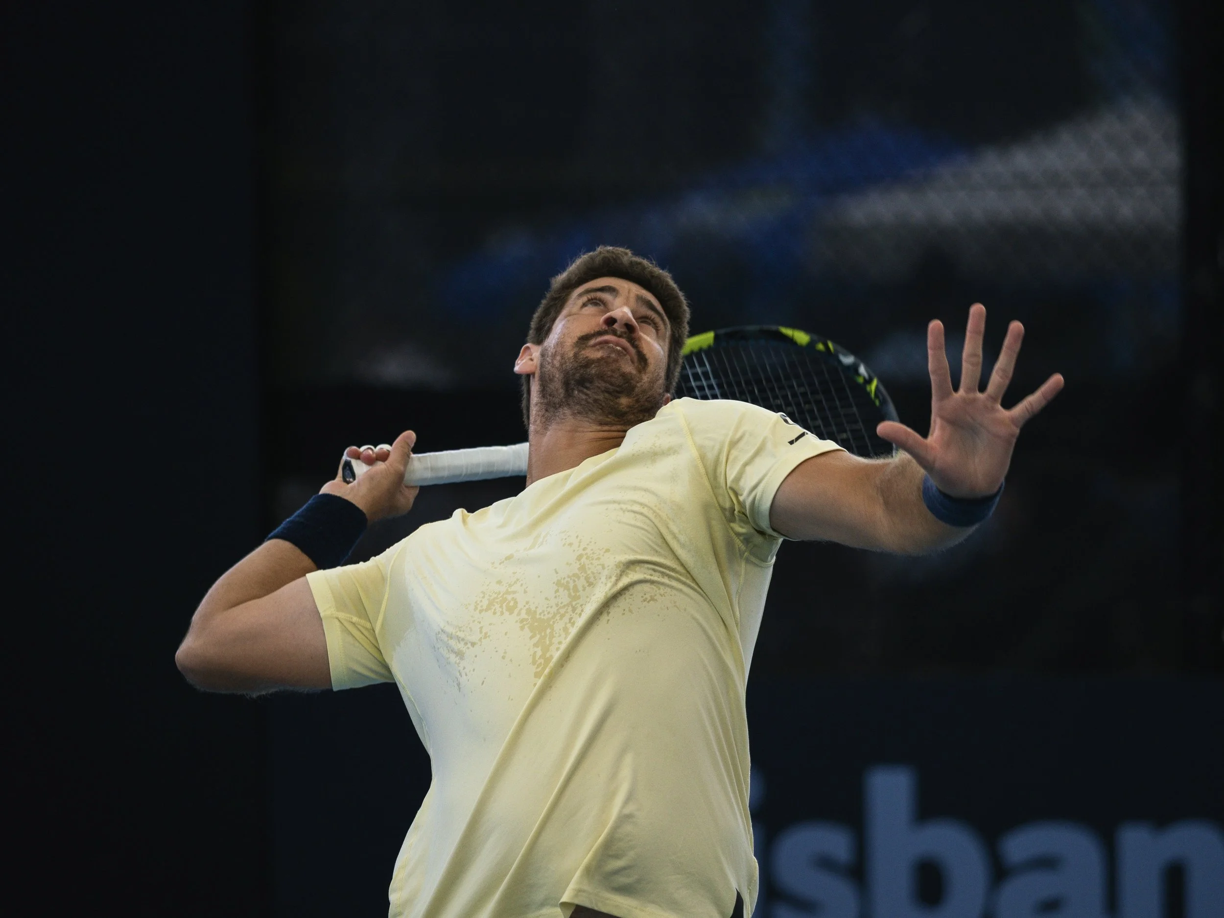 A male tennis player in a yellow shirt holding a tennis racket behind his head on a dark indoor court.