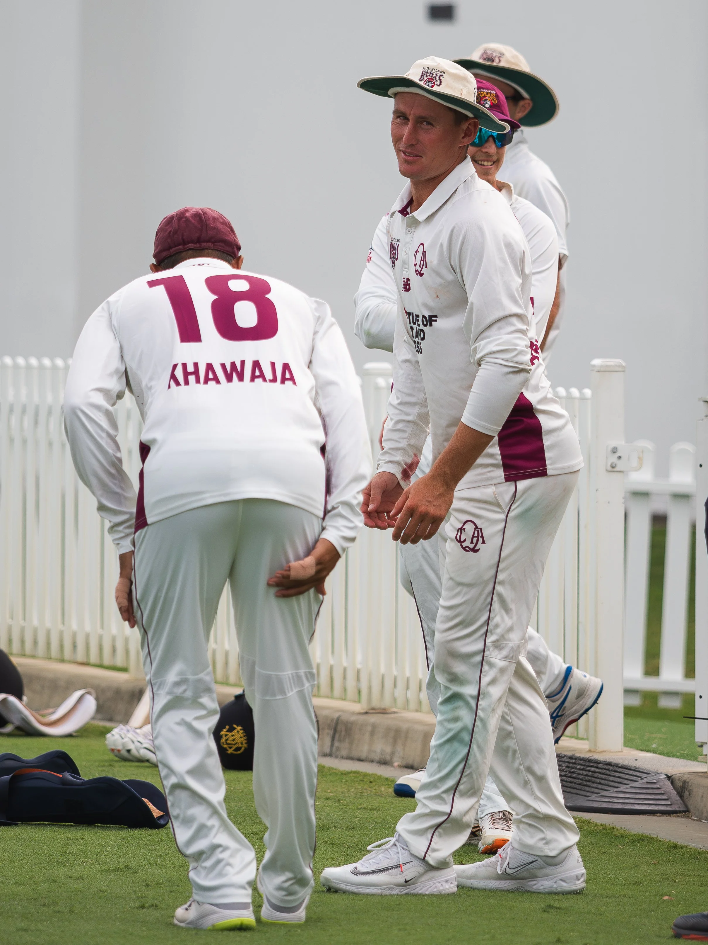 Cricket players on the field wearing white uniforms and wide-brimmed hats, in a team huddle, with equipment and a white fence in the background.